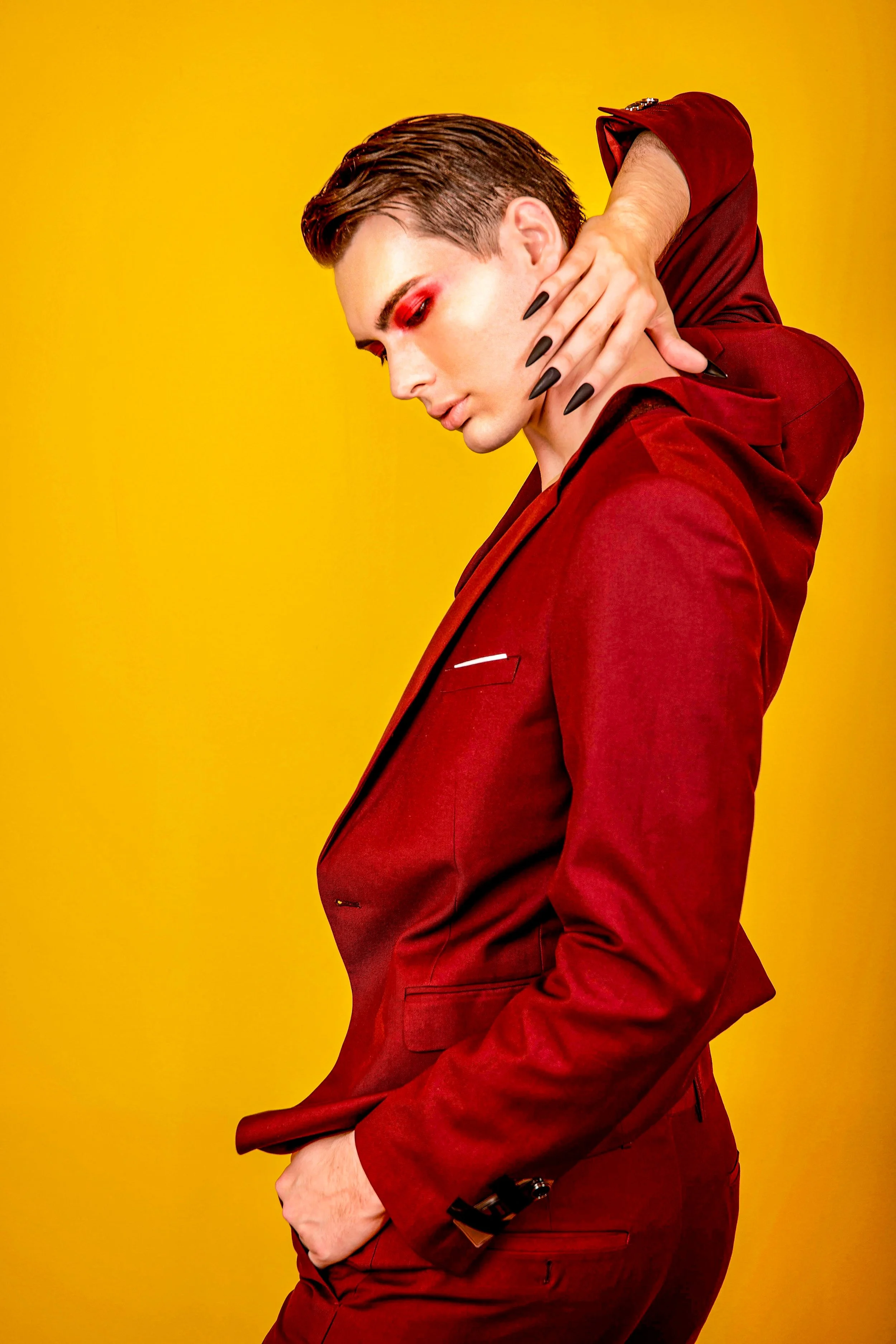 A person posed against a yellow background, wearing a red suit with matching red shirt, with short dark hair styled back, eye makeup, and black nail polish, holding their neck with one hand while the other hand is in their pocket.