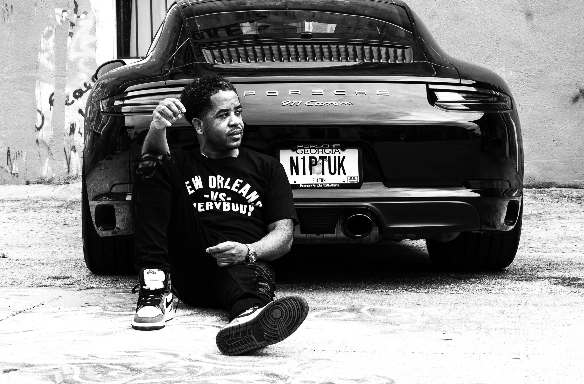A young man sitting on the ground in front of a black Porsche 911 Carrera with a Georgia license plate, wearing a black T-shirt that says 'New Orleans V. vs. Everybody' and Nike sneakers, with one shoe untied.