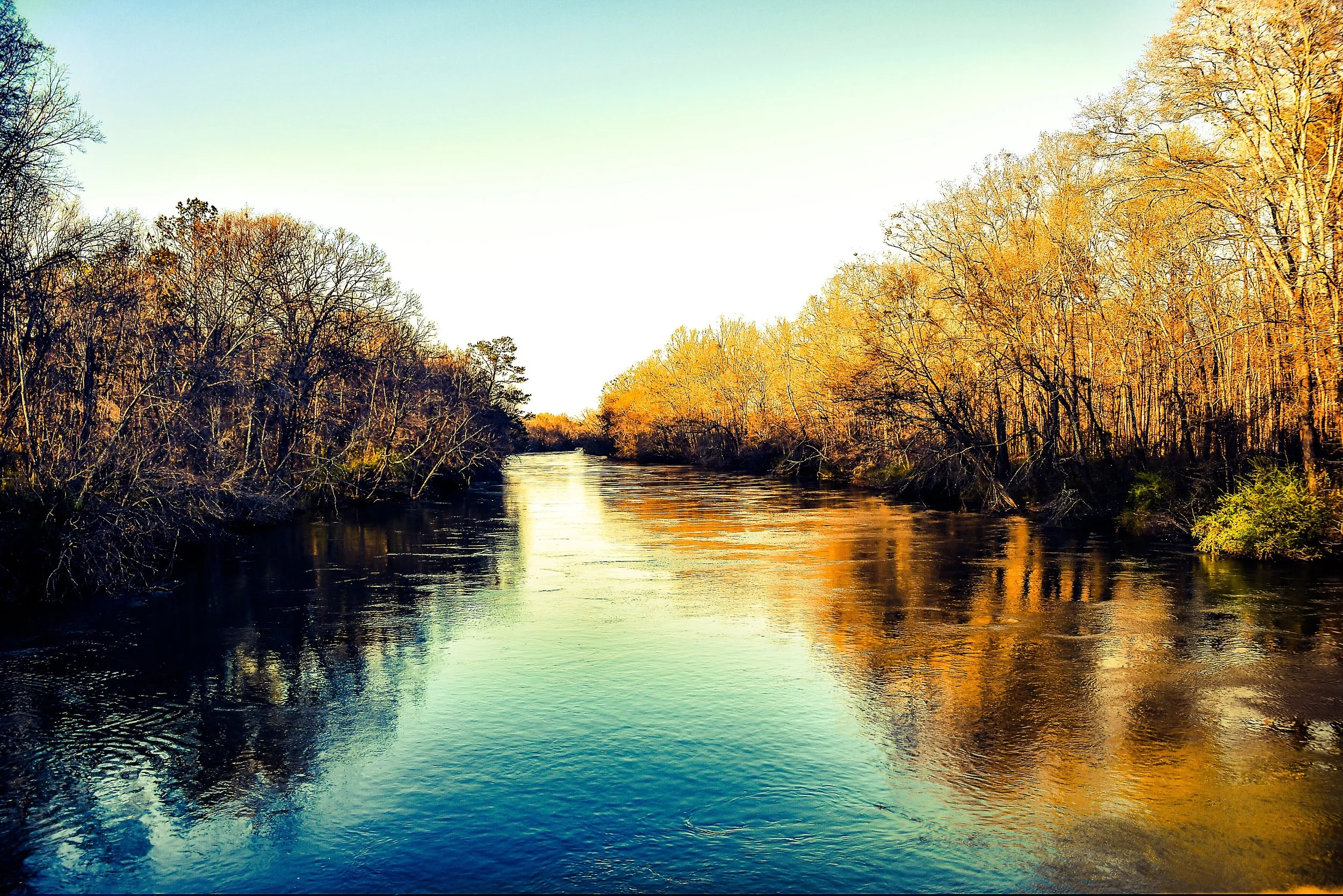 A river flowing through a wooded area with trees displaying fall foliage, under a clear sky.