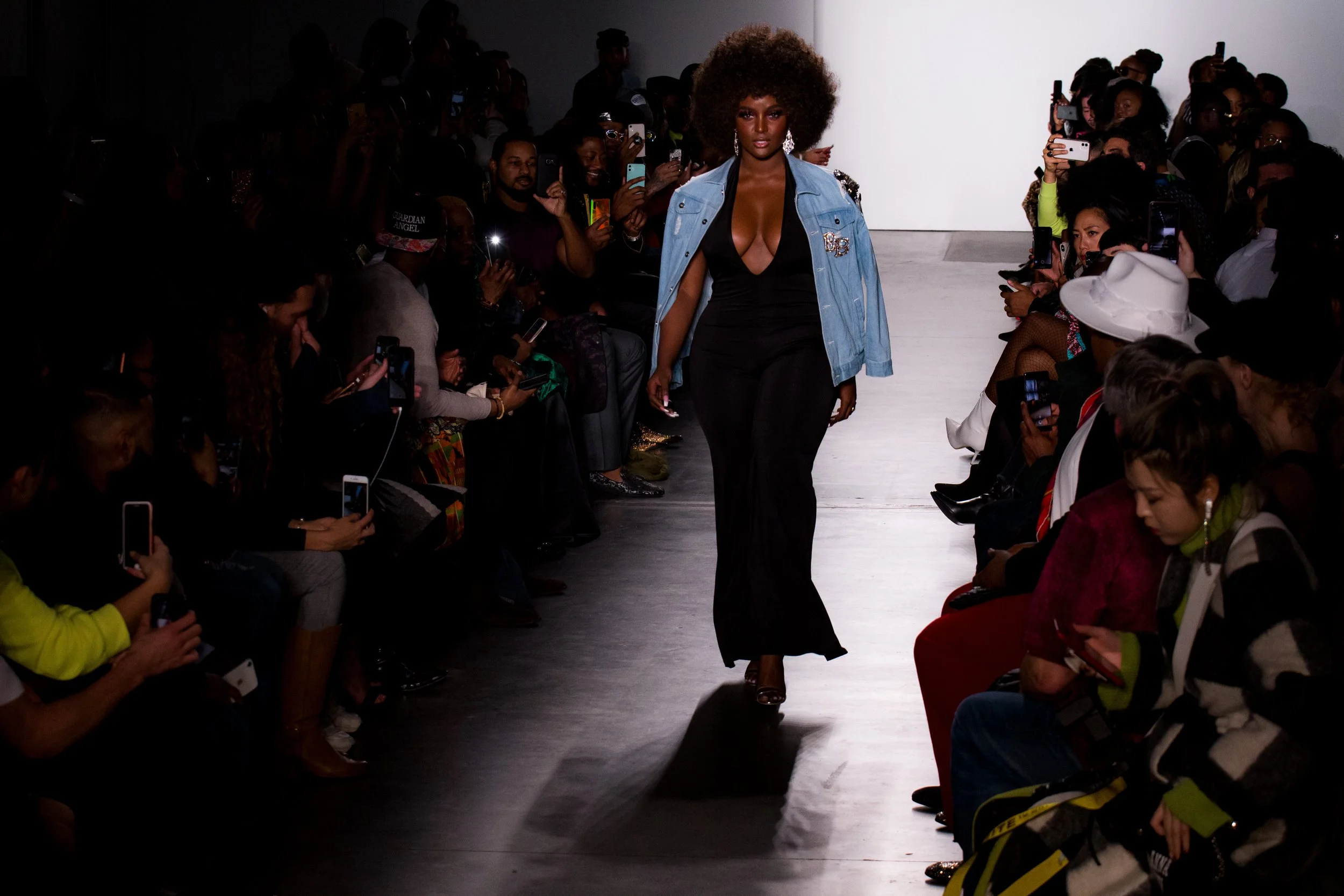 A Black female model with an Afro hairstyle walks down a fashion runway wearing a black dress with a deep neckline, a light blue denim jacket with embellishments, and high heels. The audience along both sides of the runway is taking pictures with the