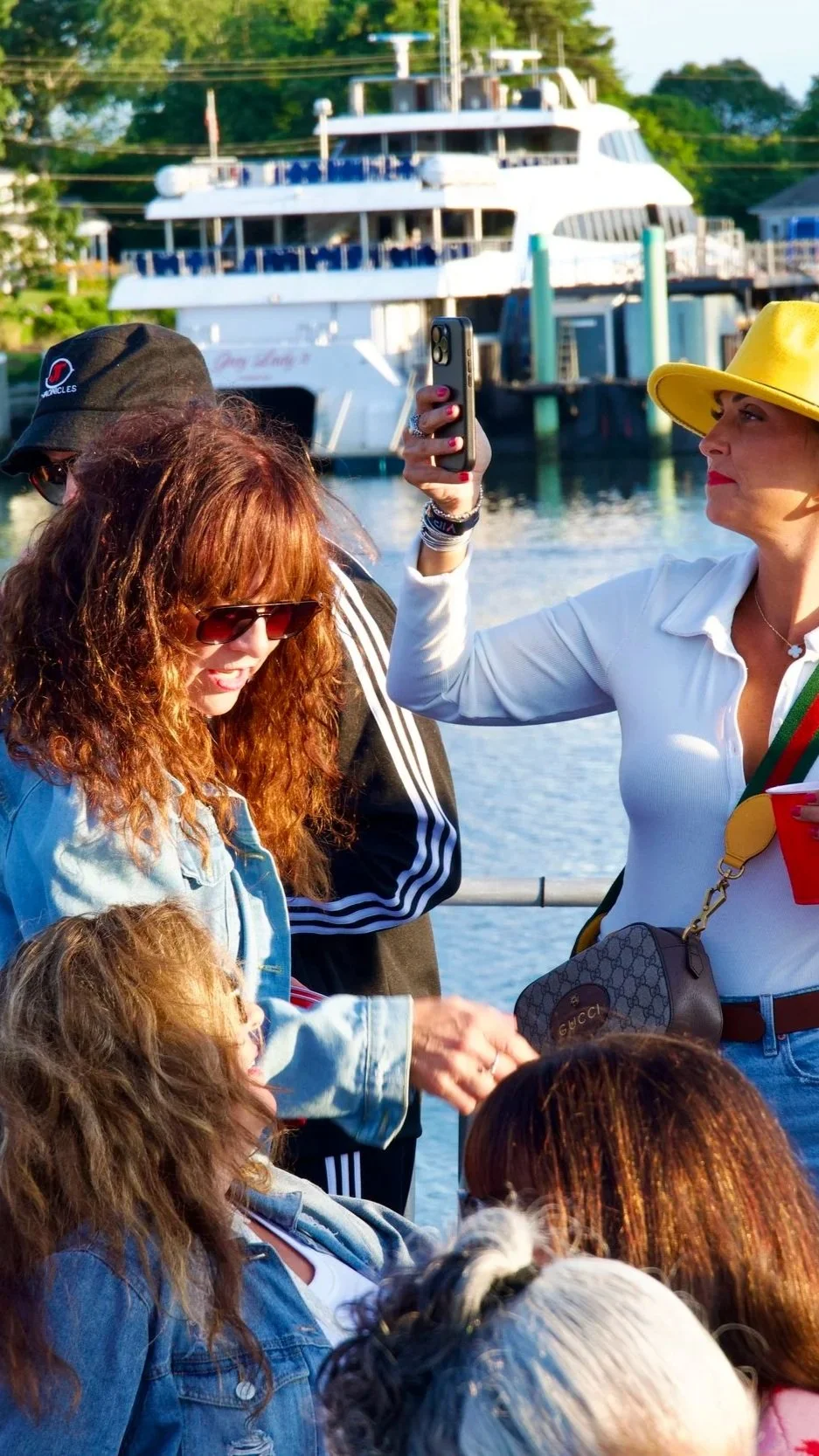 A group of women, some wearing sunglasses and hats, are gathered near a body of water with a large ferry boat in the background. One woman is taking a photo with her smartphone.