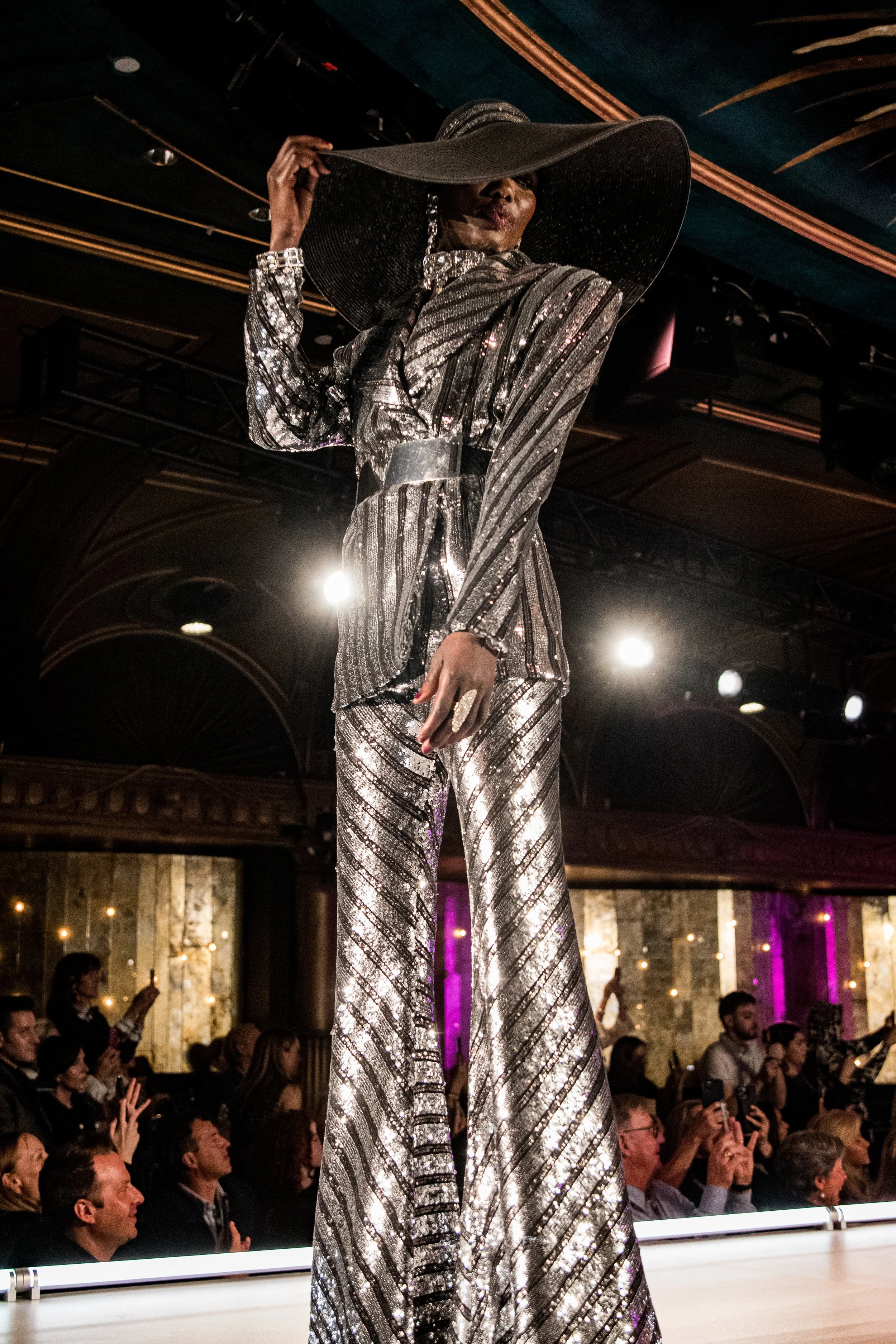 A woman on a runway wearing a large black hat and a silver metallic striped outfit with wide-leg pants, posing for the audience at a fashion show.