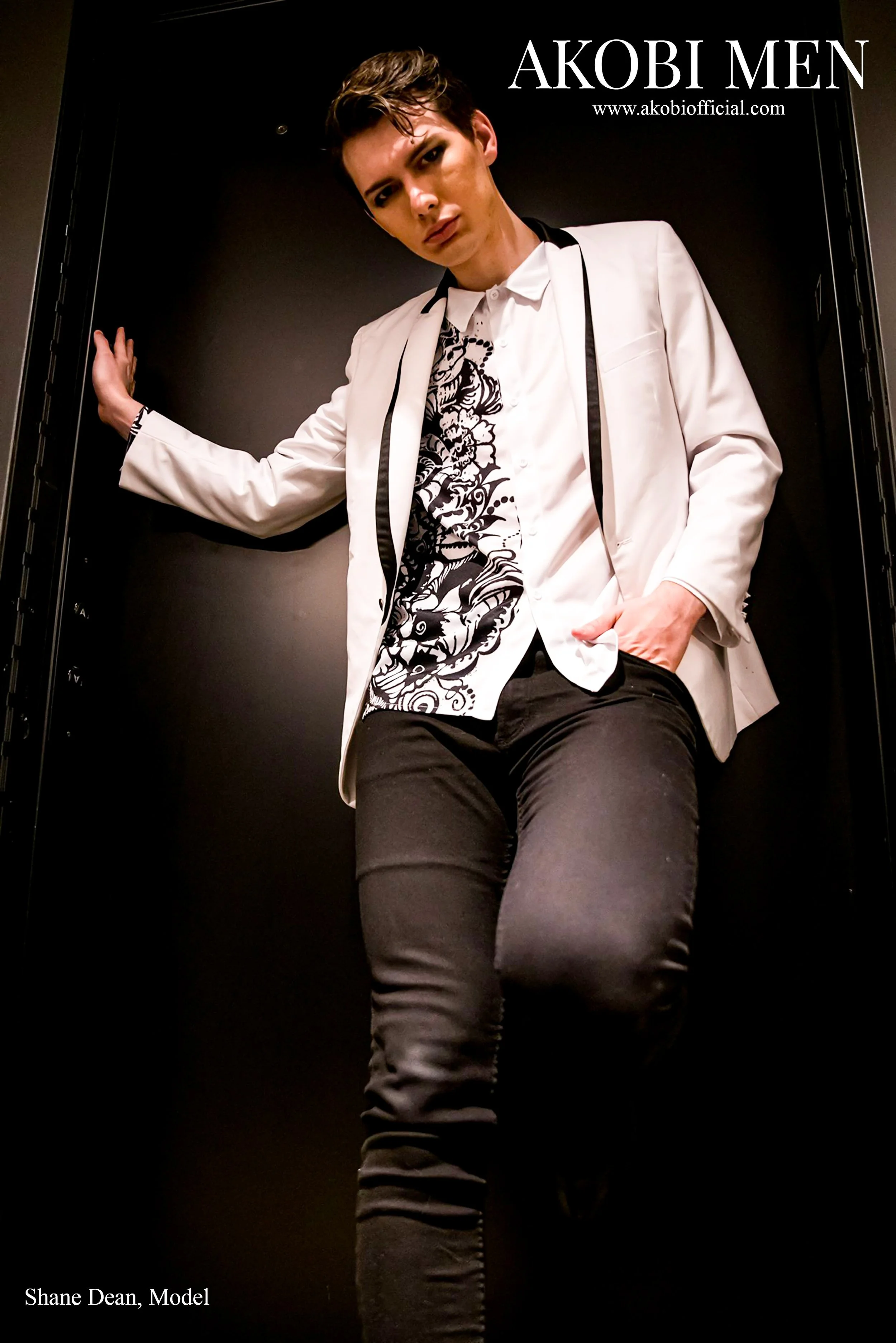 A young man posing against a black background, wearing a white blazer, a shirt with a black and white floral pattern, and black pants. The image is a promotional photo for Akobi Men, featuring Shane Dean as the model.