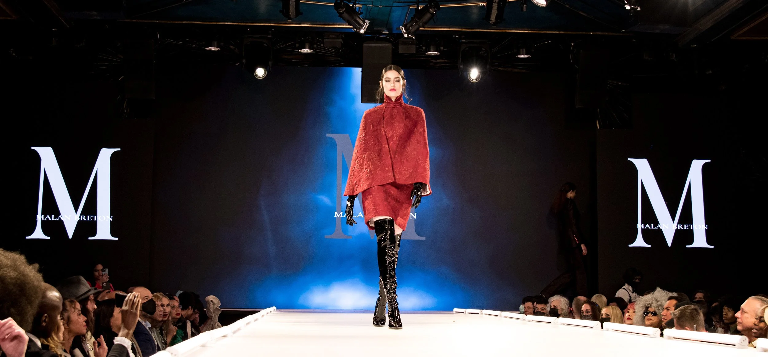 A model walking down a runway at a fashion show, wearing a red textured outfit with black glossy thigh-high boots, with an audience watching and large screens displaying a logo with the letter M and the name Malan Breton.