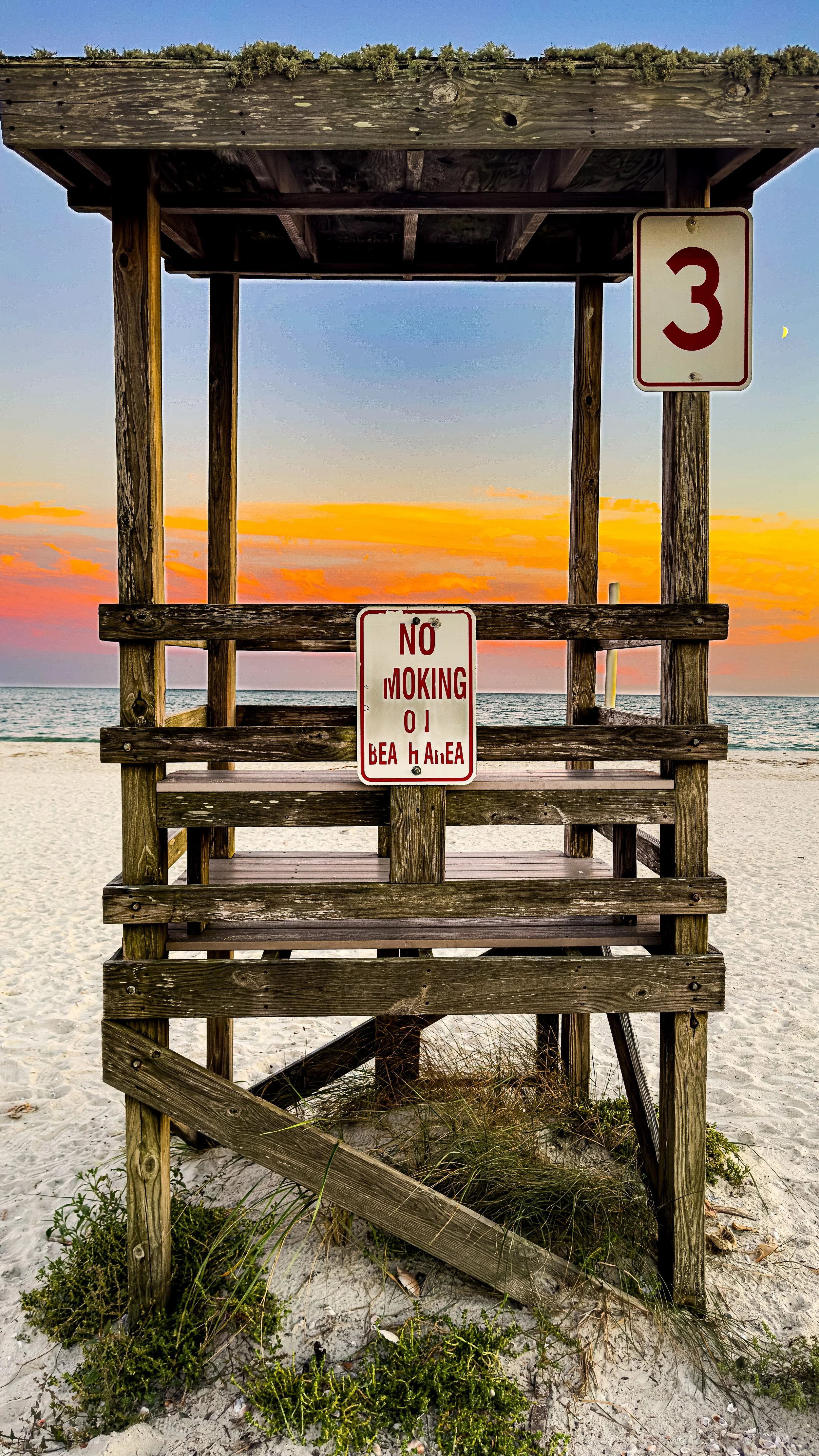 Wooden lifeguard tower on the beach with a sunset sky in the background. Sign on the tower reads 'No Moking OI Be A H Area.'