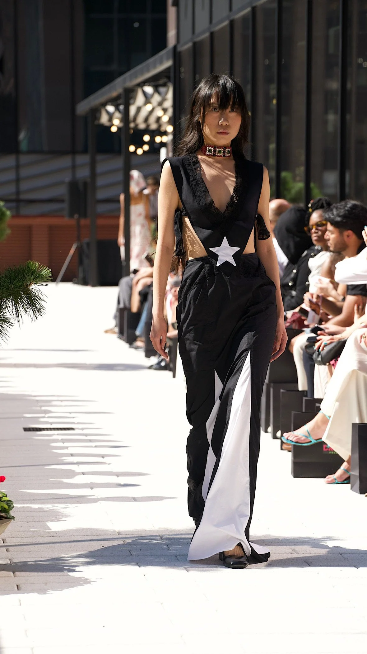 A female model walks on an outdoor runway at a fashion show, wearing a black and white stylish outfit with a star design and lace details, while audience members seated on either side watch.