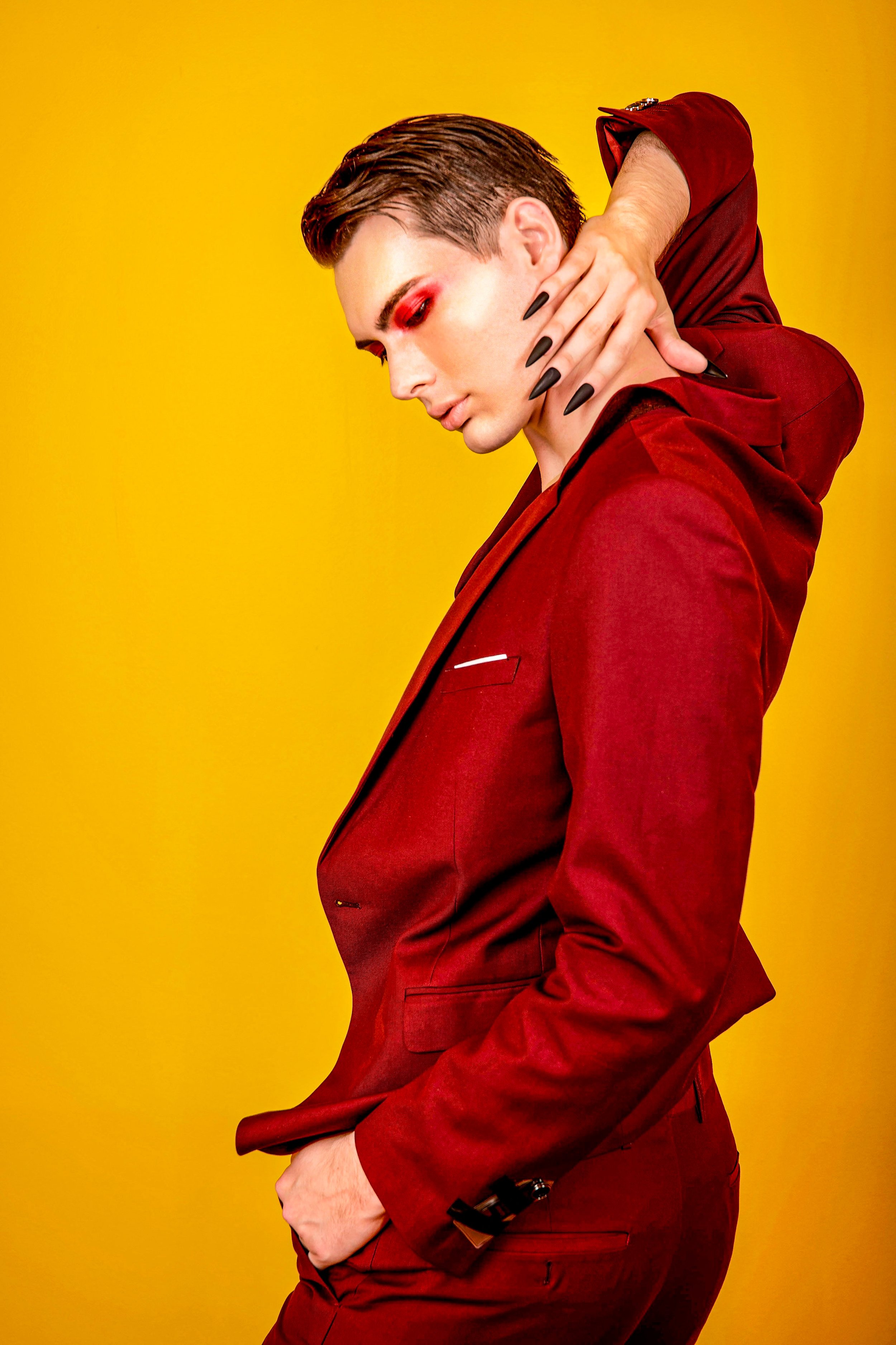 A person with short, styled hair and dramatic red eye makeup, dressed in a red suit, poses against a yellow background.