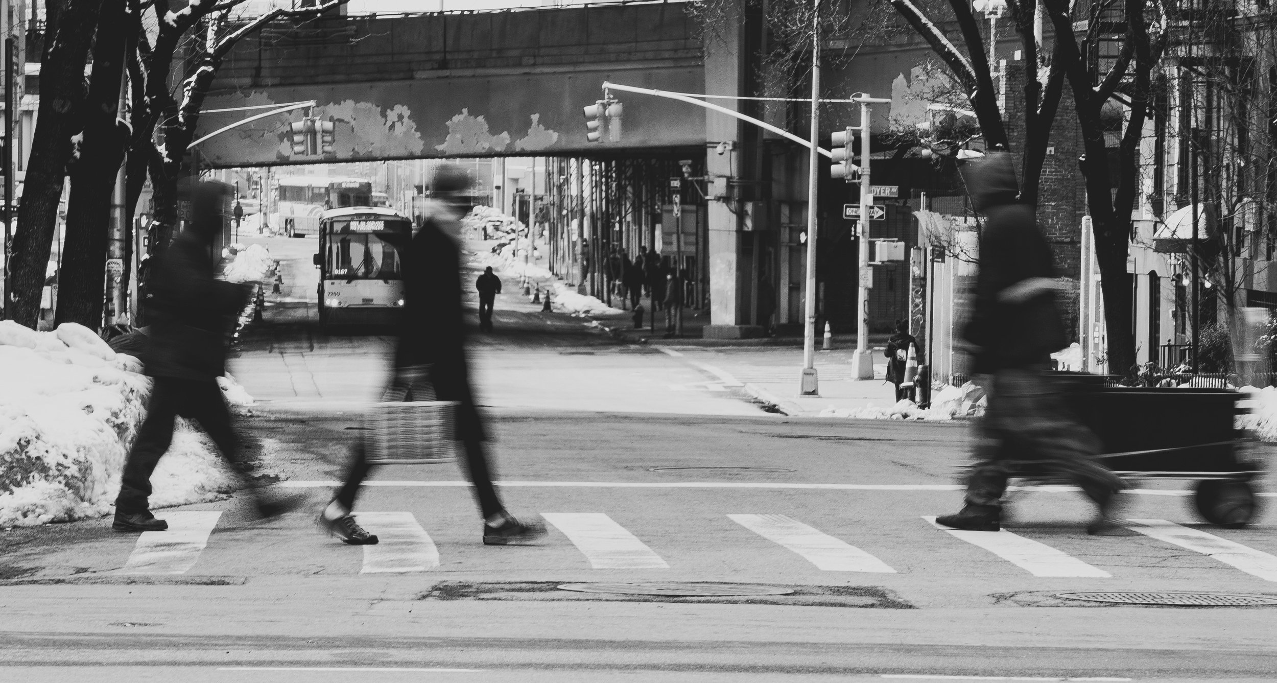 Black and white photo of a city street scene with pedestrians crossing at a crosswalk and a cyclist riding by. Snow is on the ground and trees line the street.