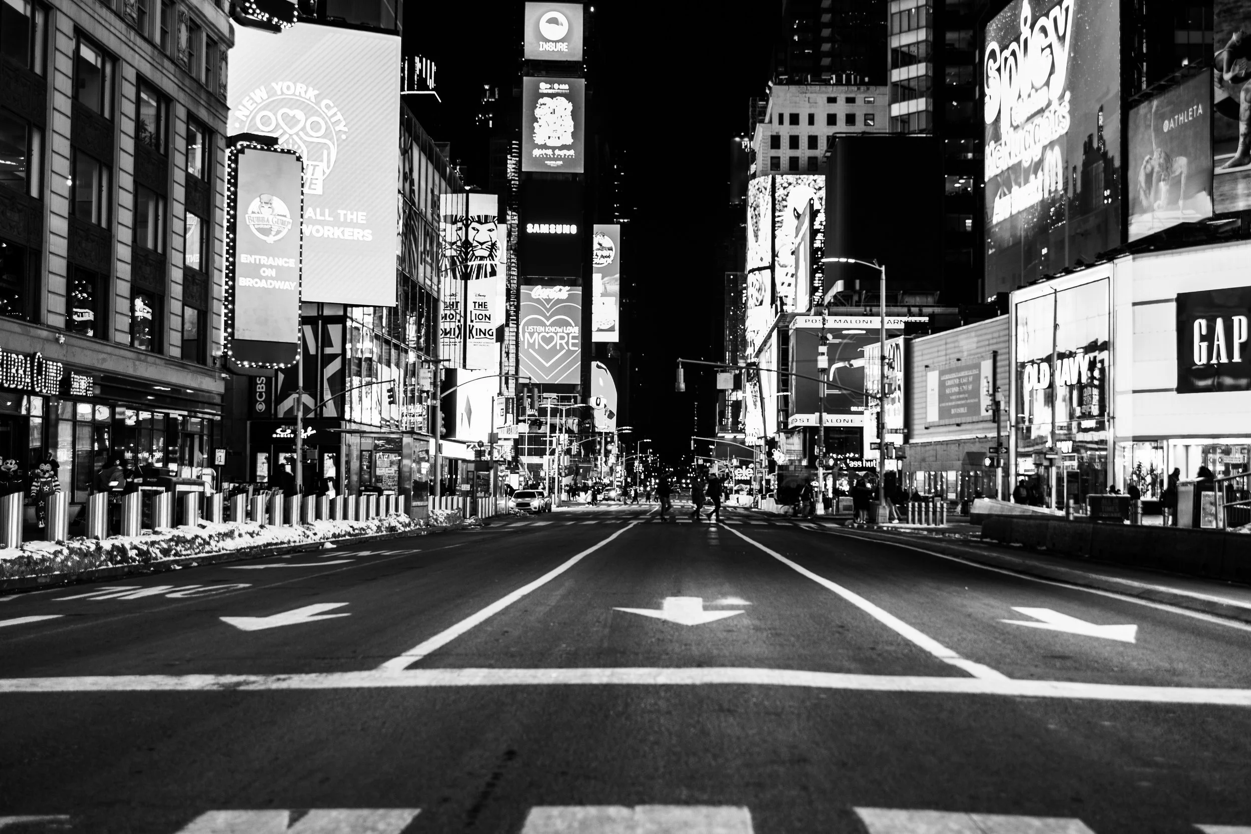 Nighttime street scene in New York City with illuminated billboards and advertisements on tall buildings, empty road with white lane markings, and some pedestrians and vehicles.