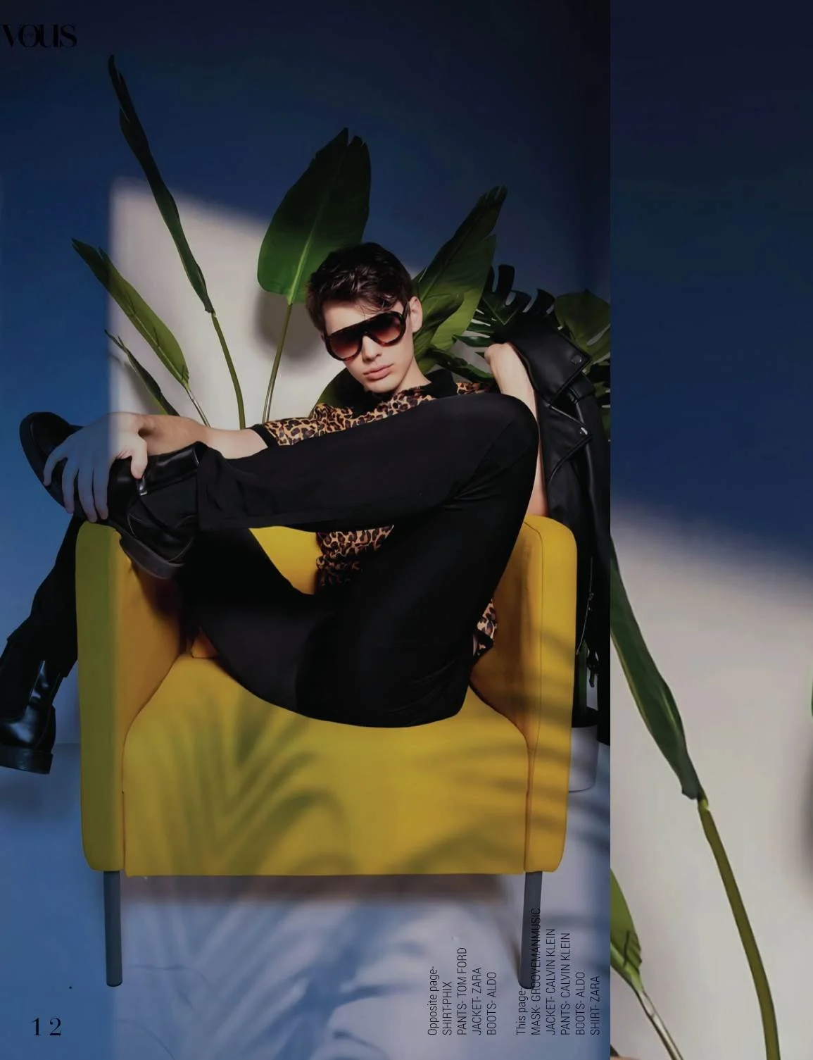A young man sitting on a yellow chair with a large green plant behind him, wearing sunglasses, a leopard print shirt, black pants, and black boots. He has a black jacket draped over his shoulder and is striking a pose.