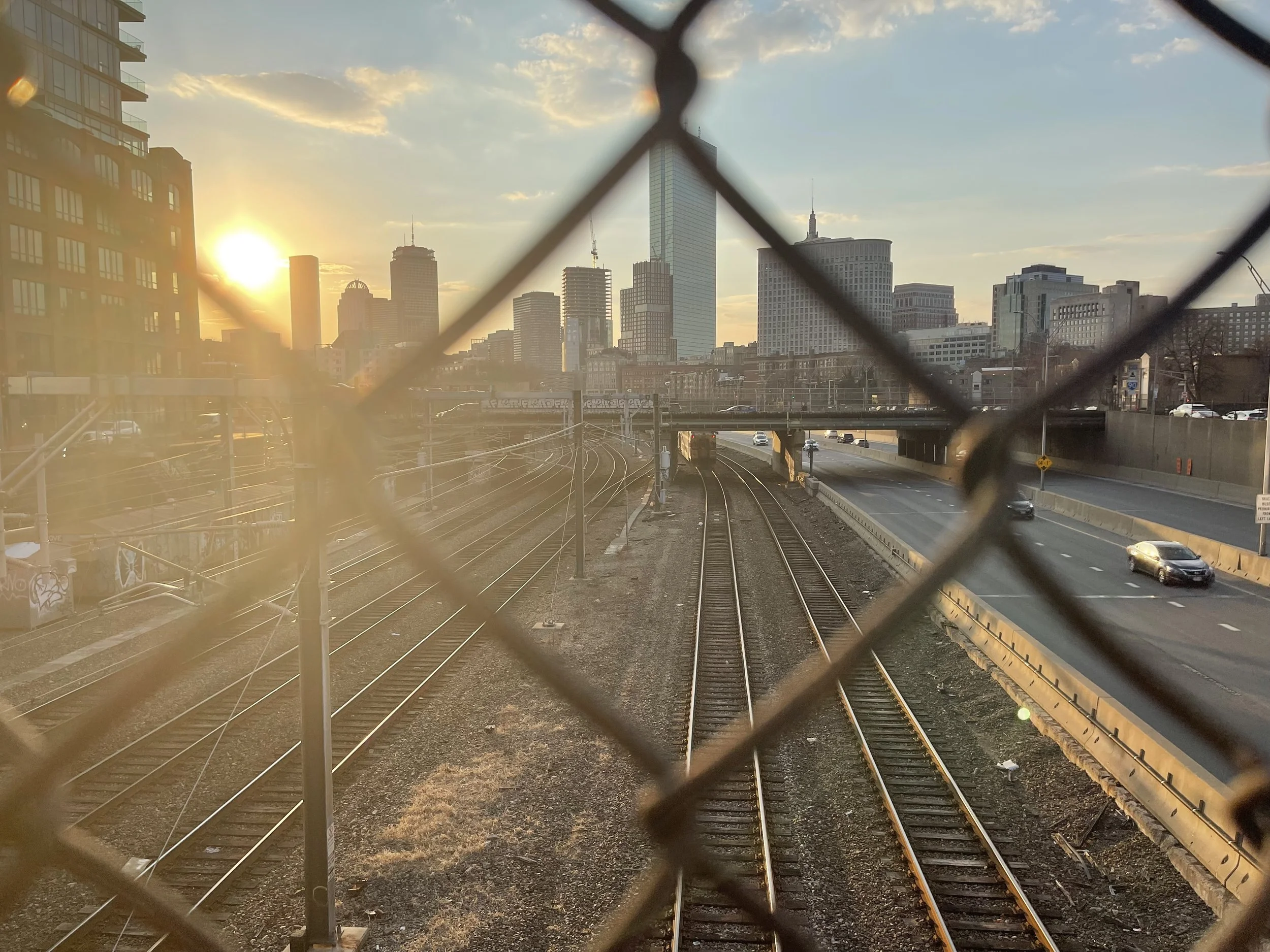 City skyline with tall buildings and skyscrapers at sunset, viewed through a chain-link fence over railroad tracks.