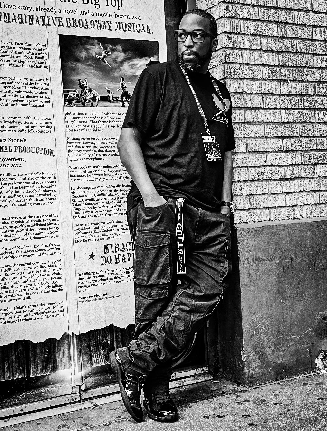 A man with glasses and a beard leaning against a brick wall and a large poster or newspaper backdrop. The man wears a black t-shirt, tactical-style pants, and boots, with a lanyard around his neck. The photo is in black and white.