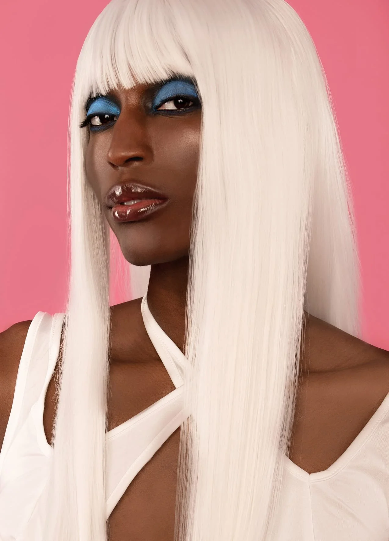 A woman with dark skin and platinum blonde hair styled long and straight, wearing bold blue eye makeup, shiny lip gloss, and a white sleeveless top, with a pink background.