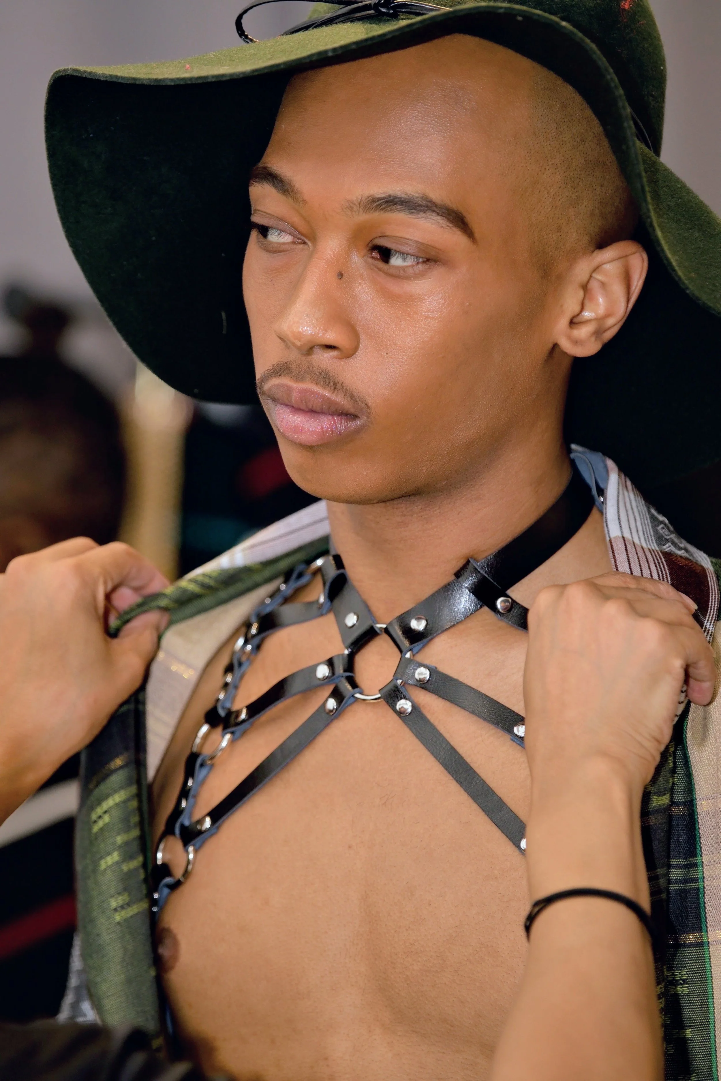 Close-up of a person with a shaved head wearing a wide-brimmed hat, with a sheet of black leather with metal rings draped around their neck, over a partially visible striped shirt, and a blurred background.
