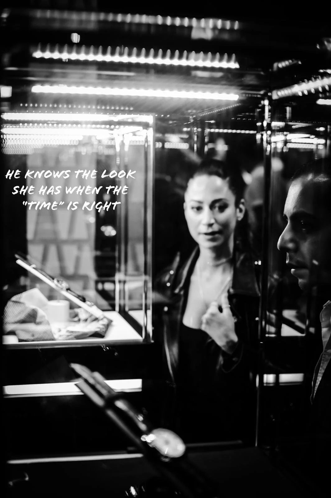 Black and white photo of a woman and a man reflected in a mirror, with a quote on the mirror that reads 'He knows the look she has when the "time" is right.' The woman is making a gesture with her hand on her chest, and both are dressed in dark cloth