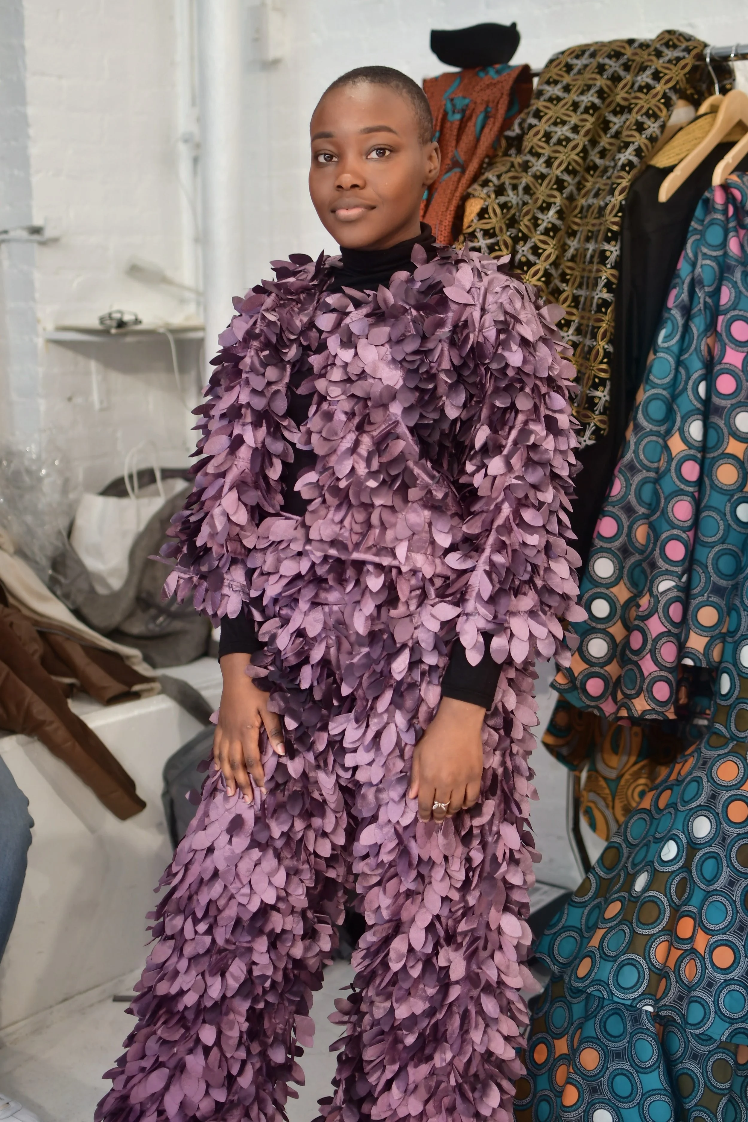 A woman standing in front of a clothing rack, wearing a dress made of numerous purple leaves.