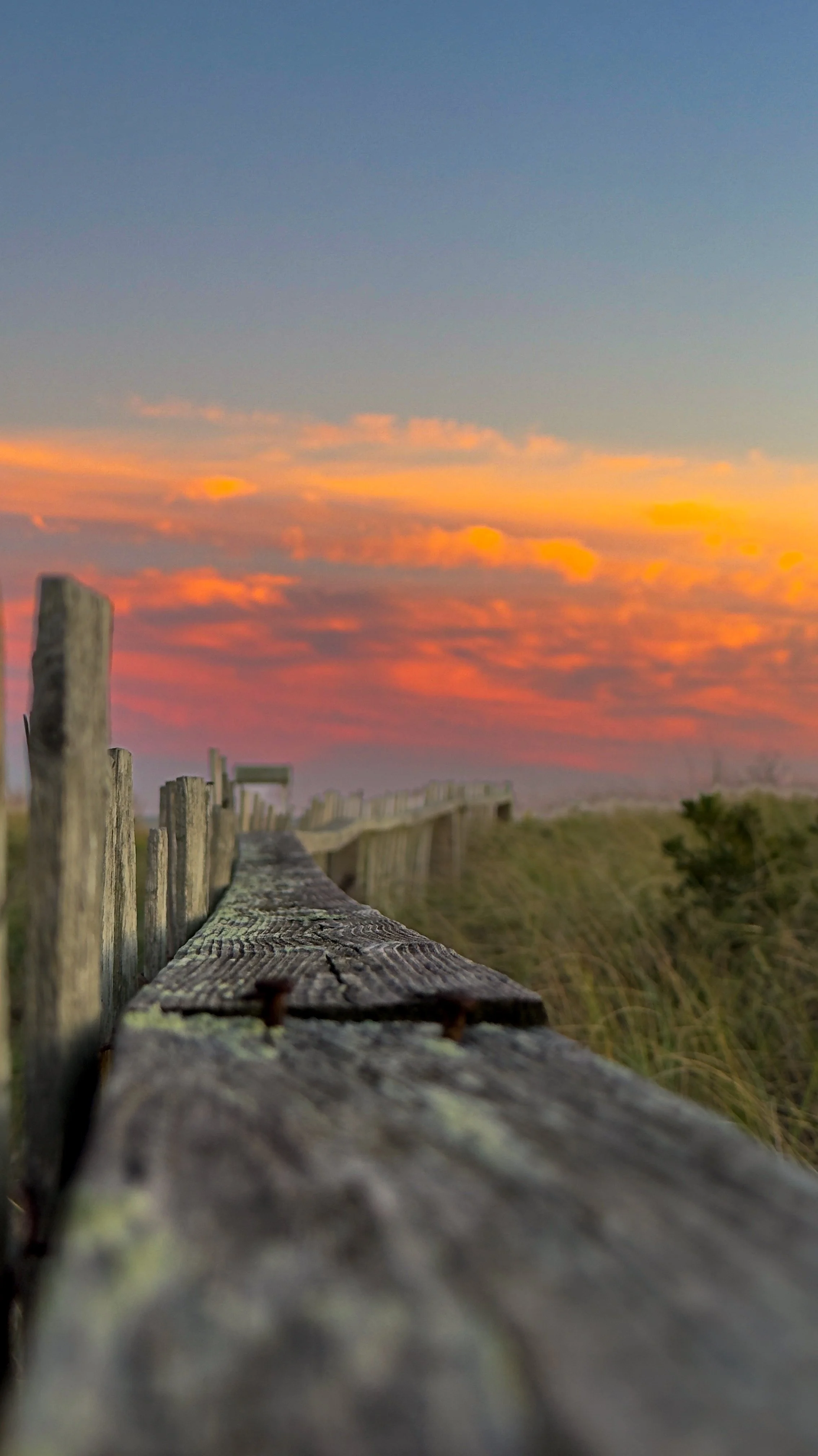 A close-up of a weathered wooden fence along a sandy path at sunset, with a sky filled with colorful orange, pink, and blue clouds in the background.