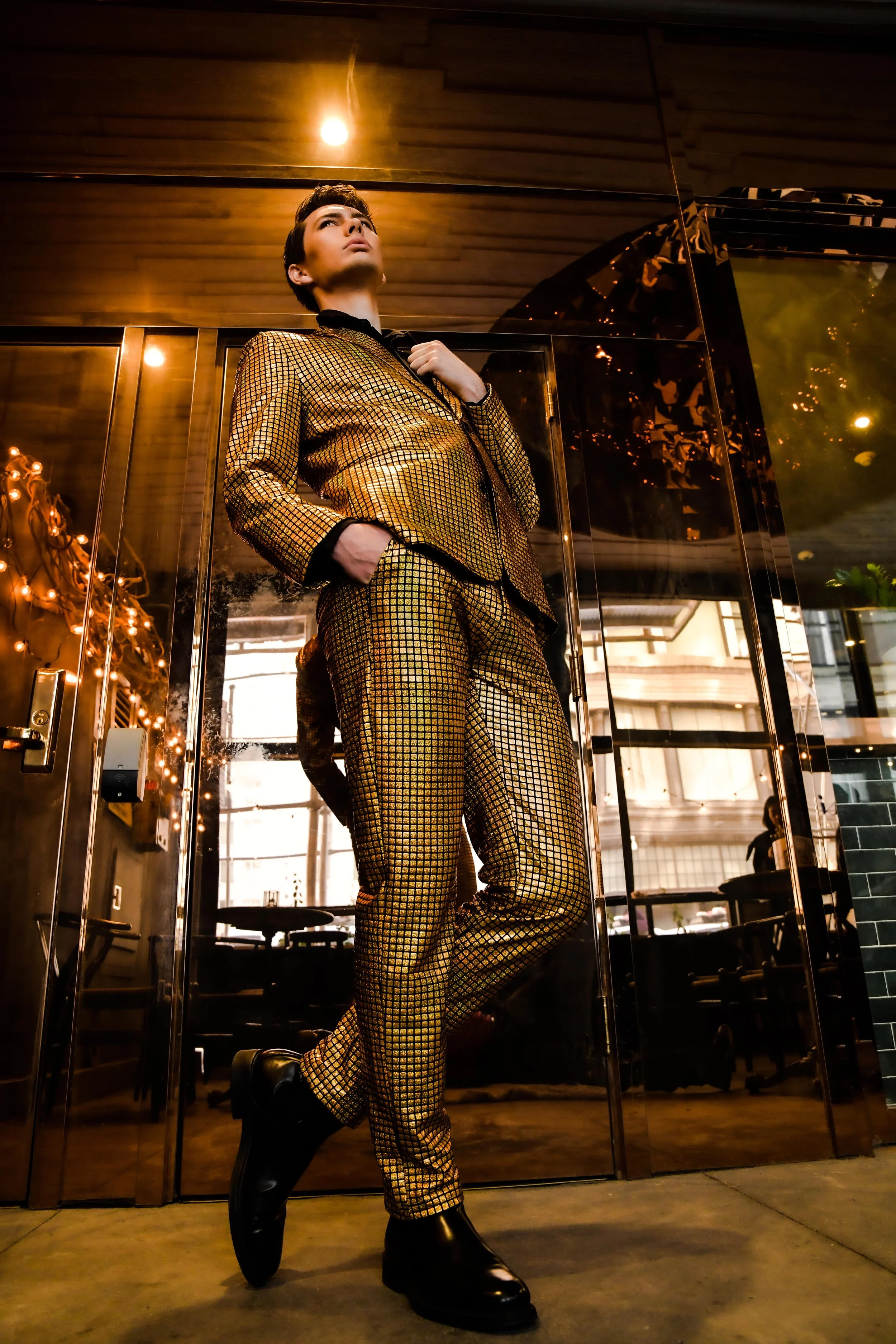 Person wearing a shiny, metallic gold and black checkered suit, standing inside a modern building with glass doors and warm lighting.