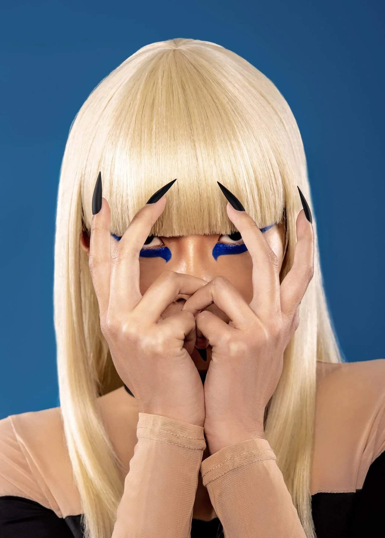 A person with platinum blonde hair and blunt bangs covering their eyes, wearing black eyeliner and nude long-sleeve gloves, makes a frame with their fingers around their eyes against a solid blue background.
