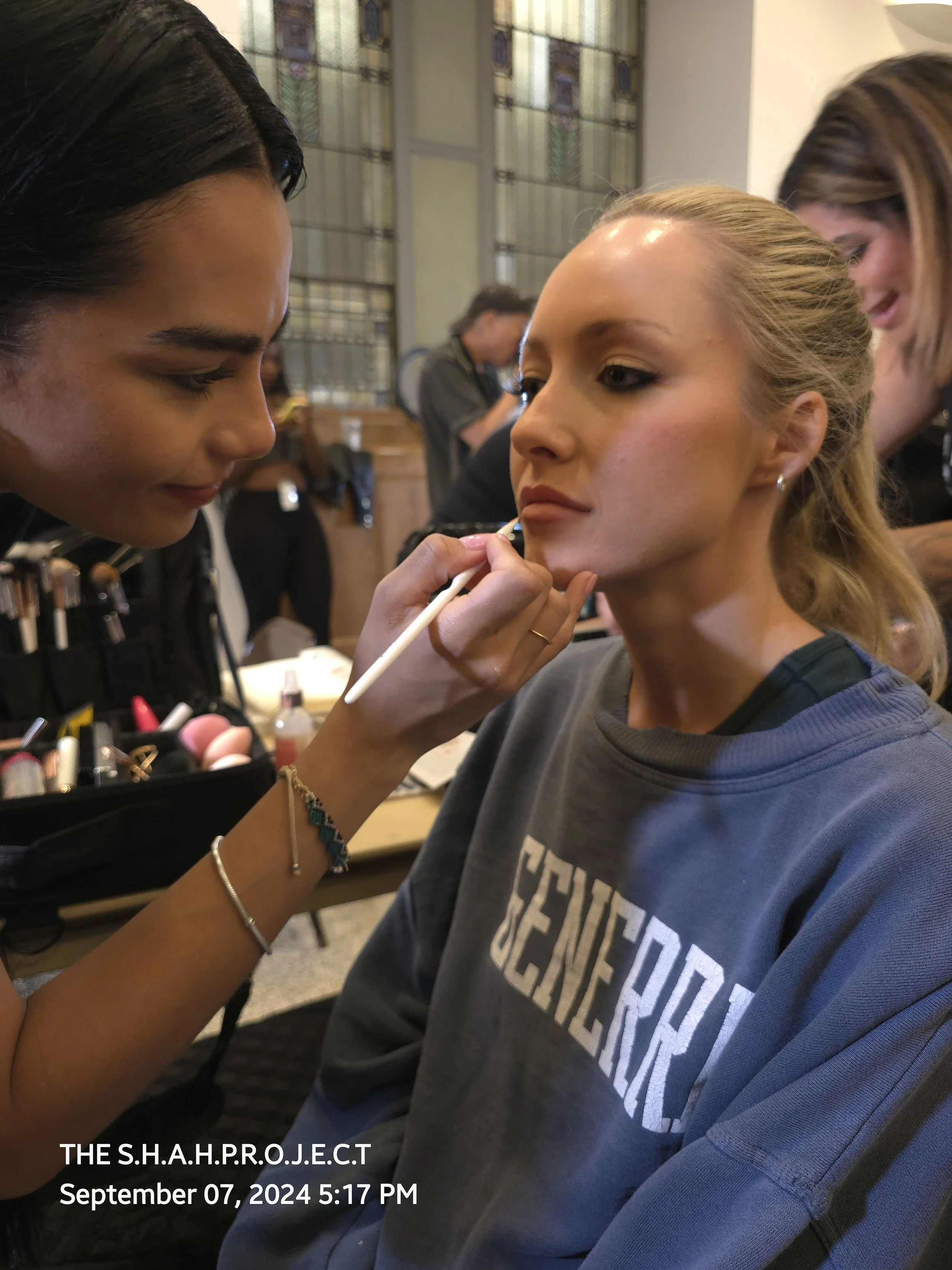 Woman in gray sweatshirt getting her makeup done by makeup artist in professional setting with stained glass window in background.
