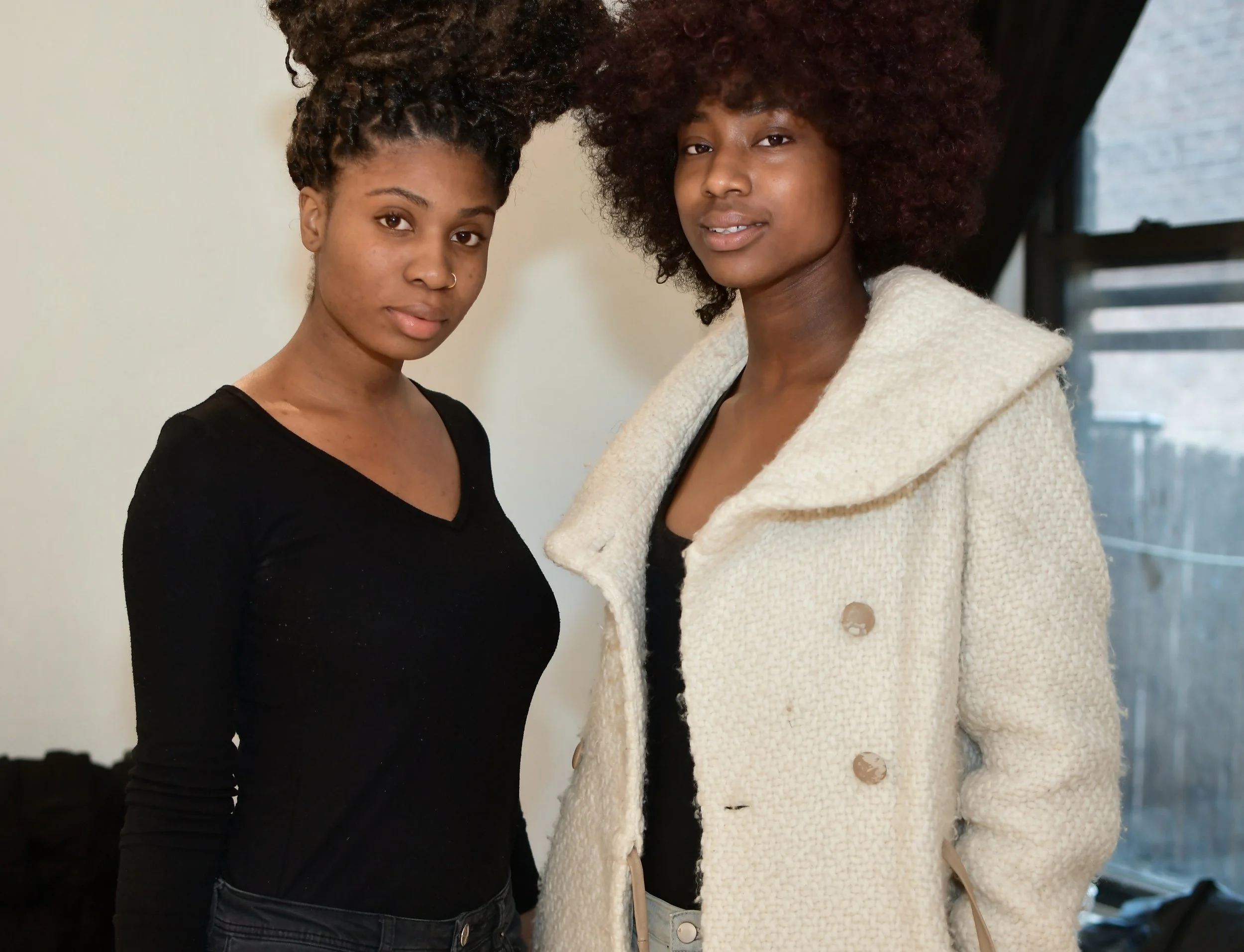 Two women with natural hair pose together indoors, one with voluminous curly hair and the other with a natural afro hairstyle, both wearing black tops and standing close in front of a plain wall and a window.