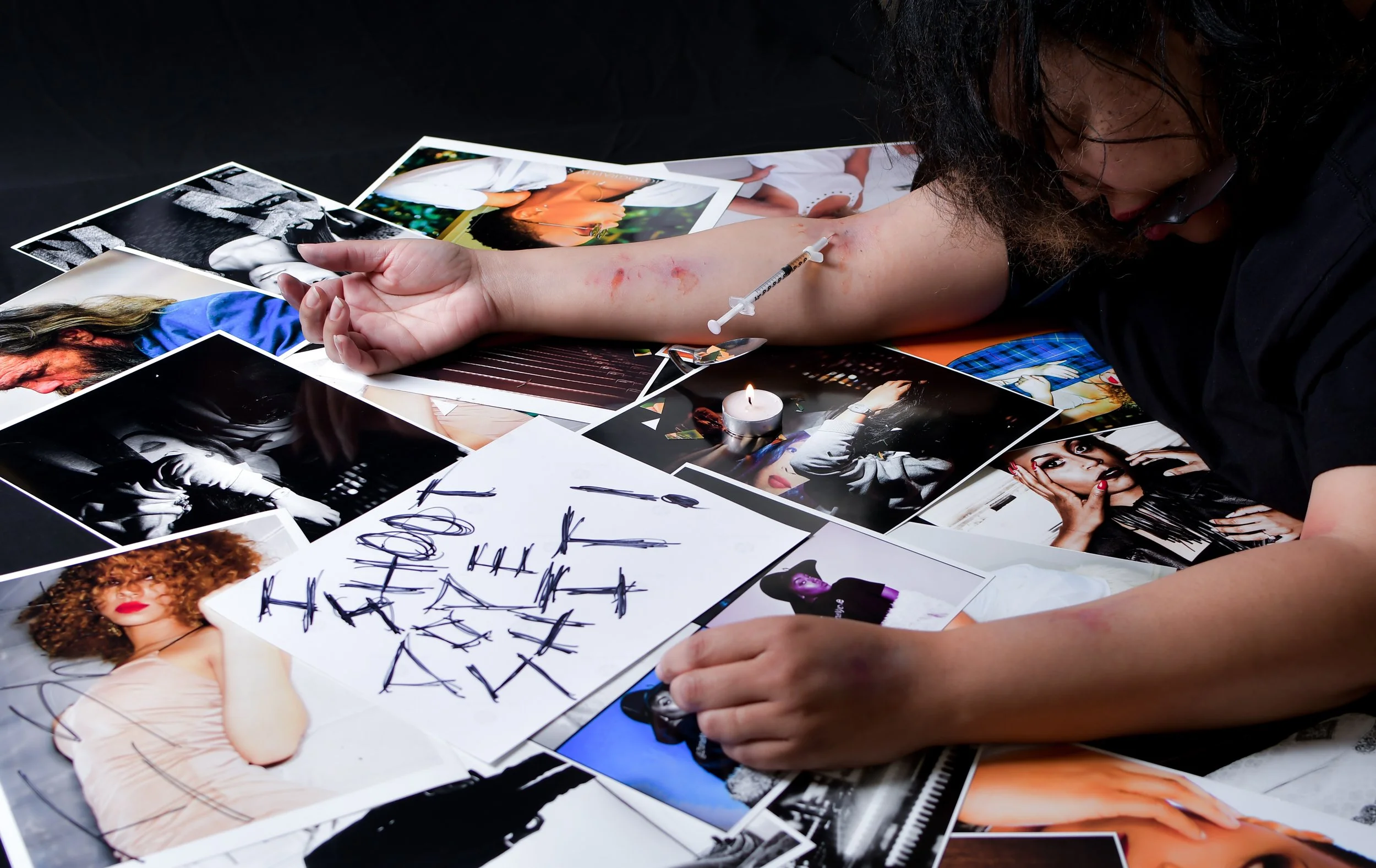 Person with black hair and glasses drawing on their arm with a syringe while surrounded by various photographs of models.