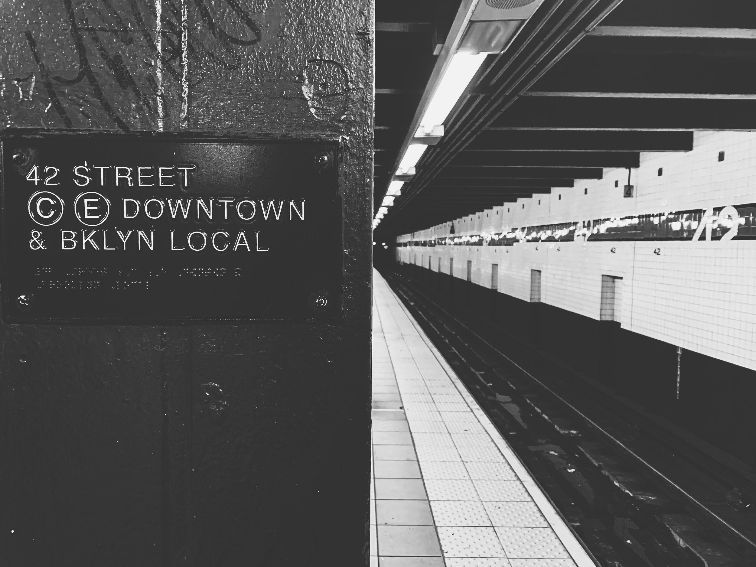 A black-and-white photo capturing an underground subway platform with a textured wall in the foreground displaying a street sign for 42nd Street in Downtown Brooklyn. The platform has a tiled floor, illuminated ceiling fixtures, and a row of empty tr
