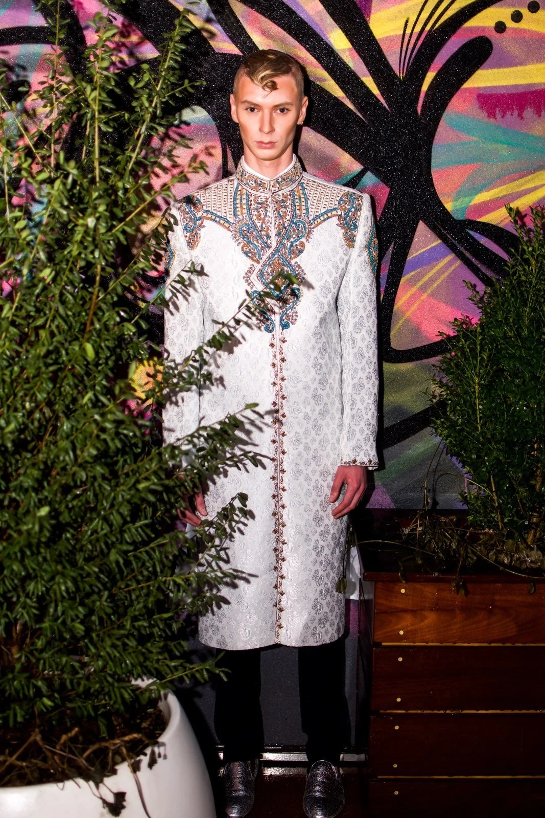 A young man standing behind shrubs with a colorful, abstract graffiti background. He is wearing a long, ornate white coat with intricate embroidery and has short, styled hair.