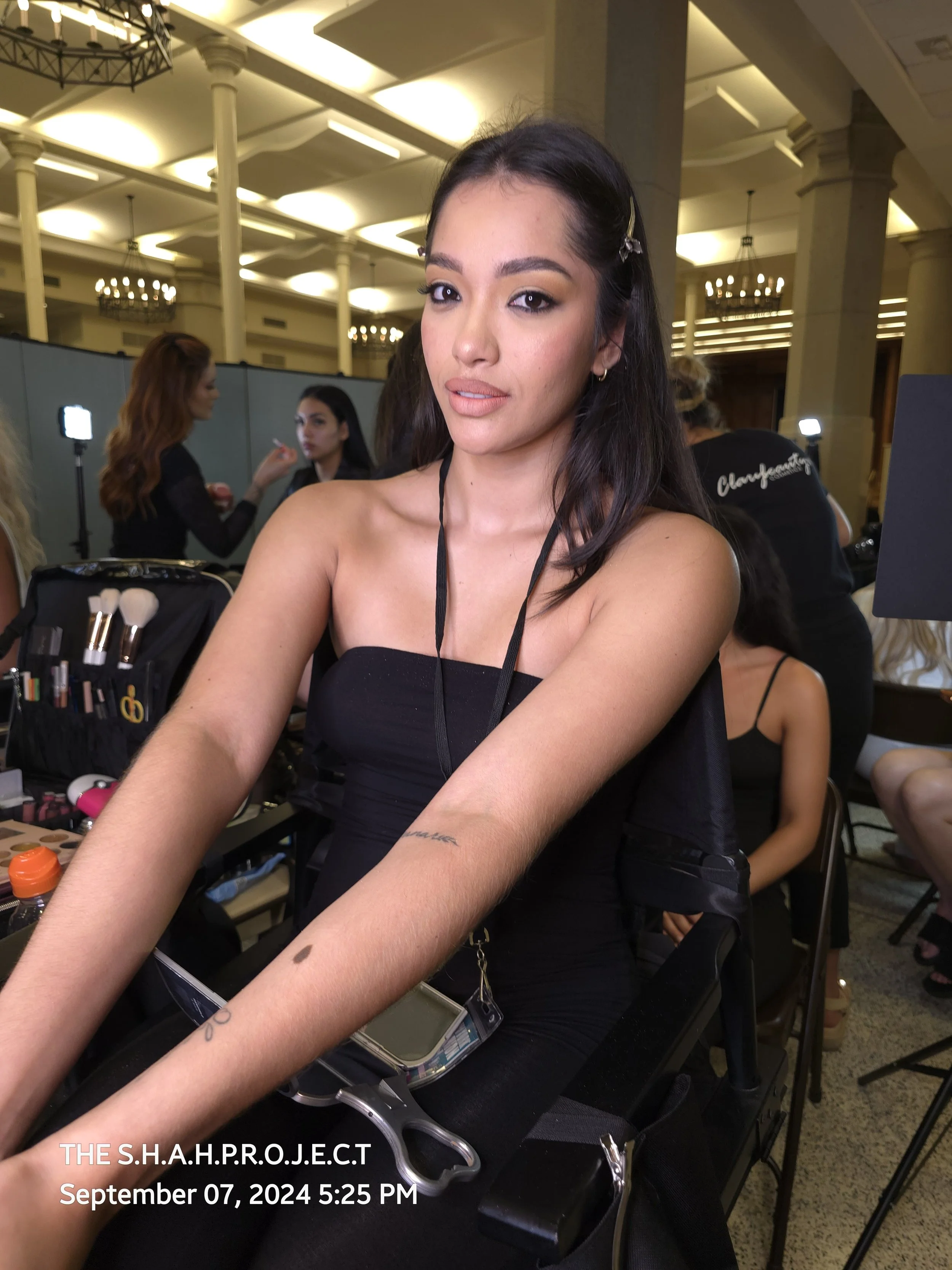 A woman in a black strapless dress sitting at a table having her makeup done by others in a hotel conference room. The room has ornate chandeliers and high ceilings.