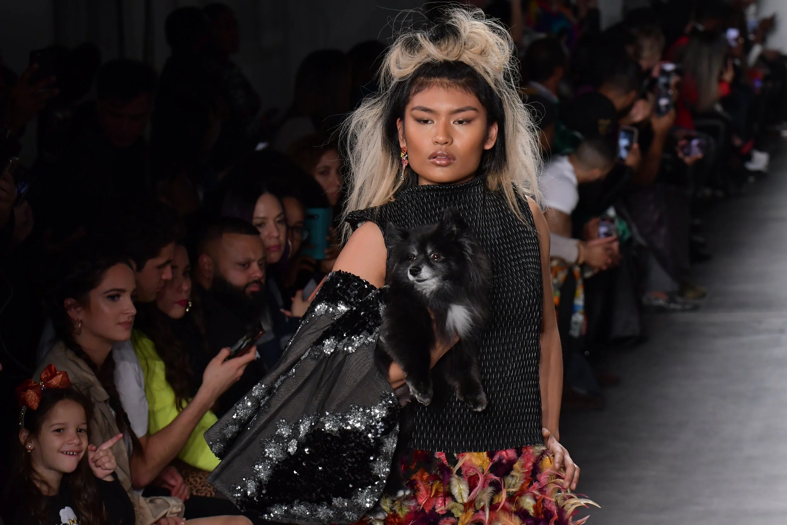 A fashion model walking down the runway during a fashion show, holding a black dog in her arms, with an audience watching in the background.
