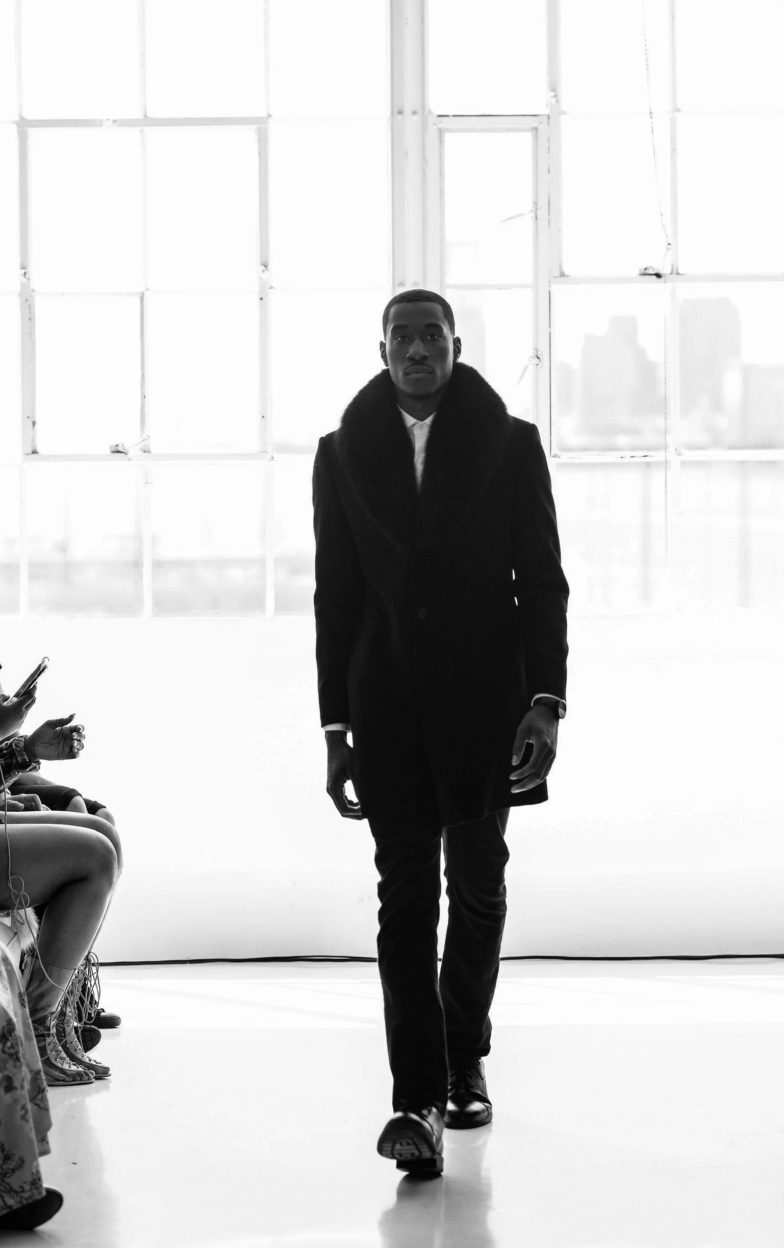 Man walking down runway at fashion show, wearing dark coat with fur collar, in front of large window with cityscape background, audience seated on side.