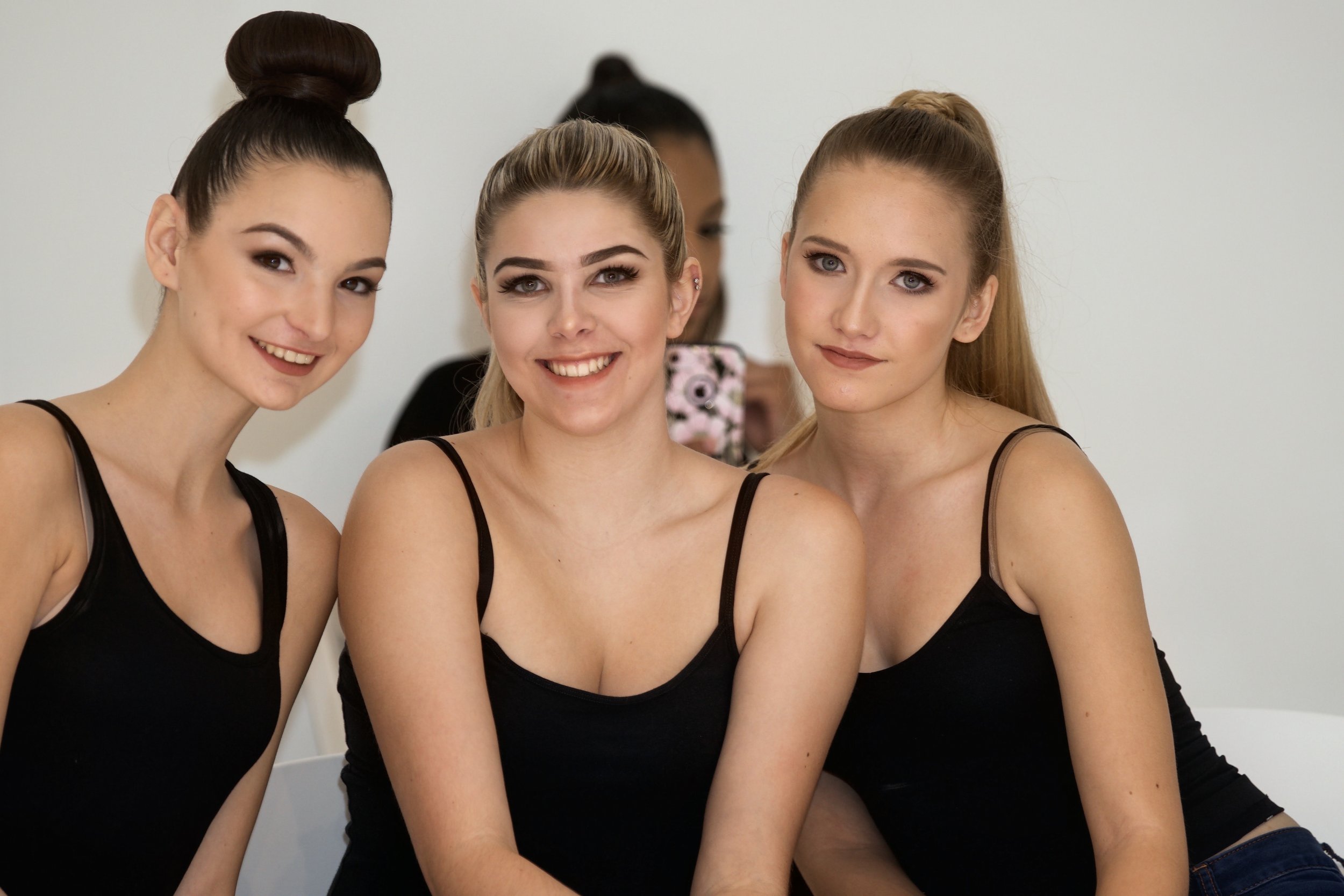 Four women sitting close together, dressed in black tank tops, smiling and looking at the camera, against a plain white background.