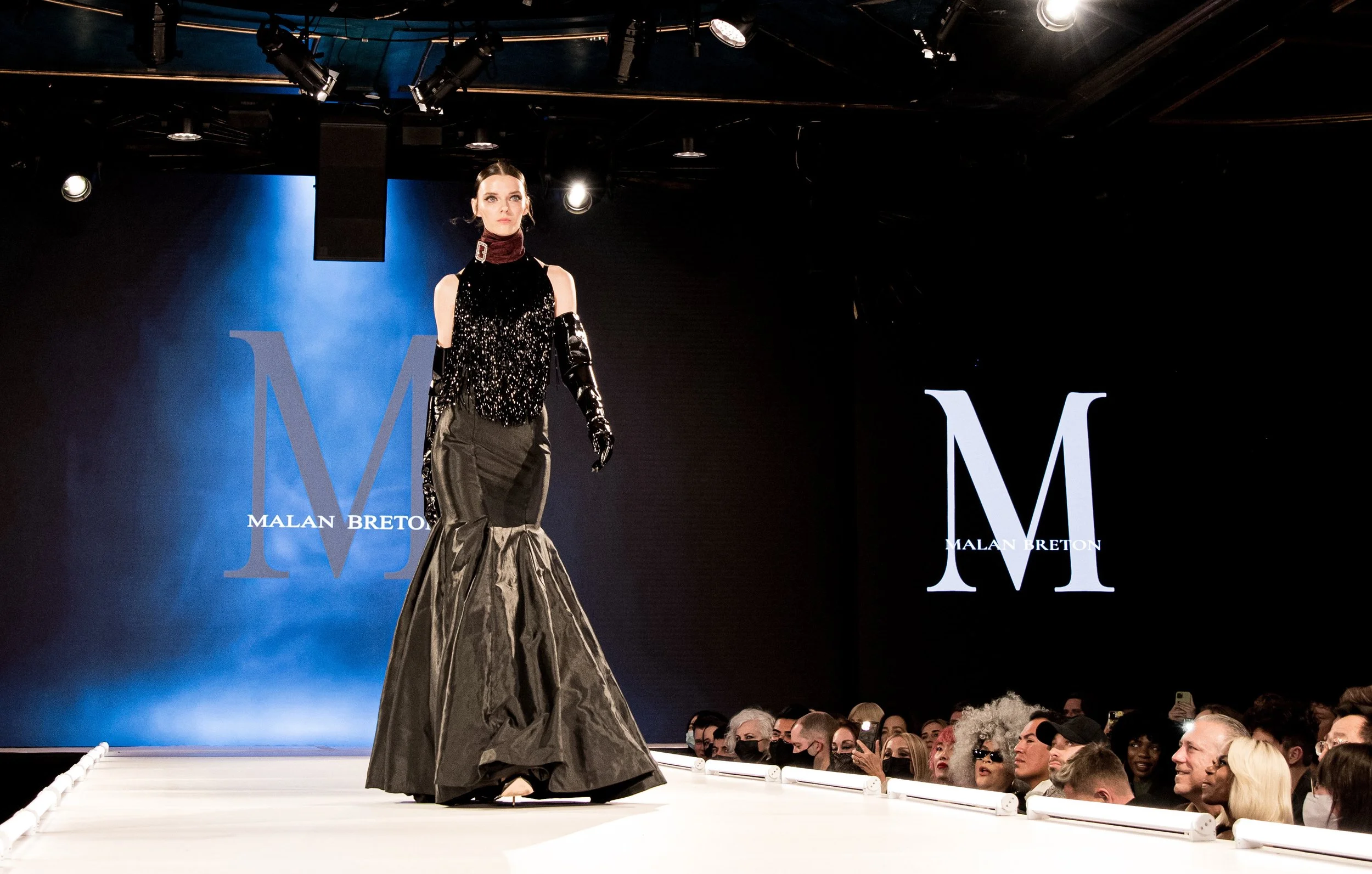 A model walking on a runway during a fashion show, wearing a black sequined top with a high collar and a long, metallic, pleated skirt. The background displays the designer's name, Malan Breton, and large initials M.