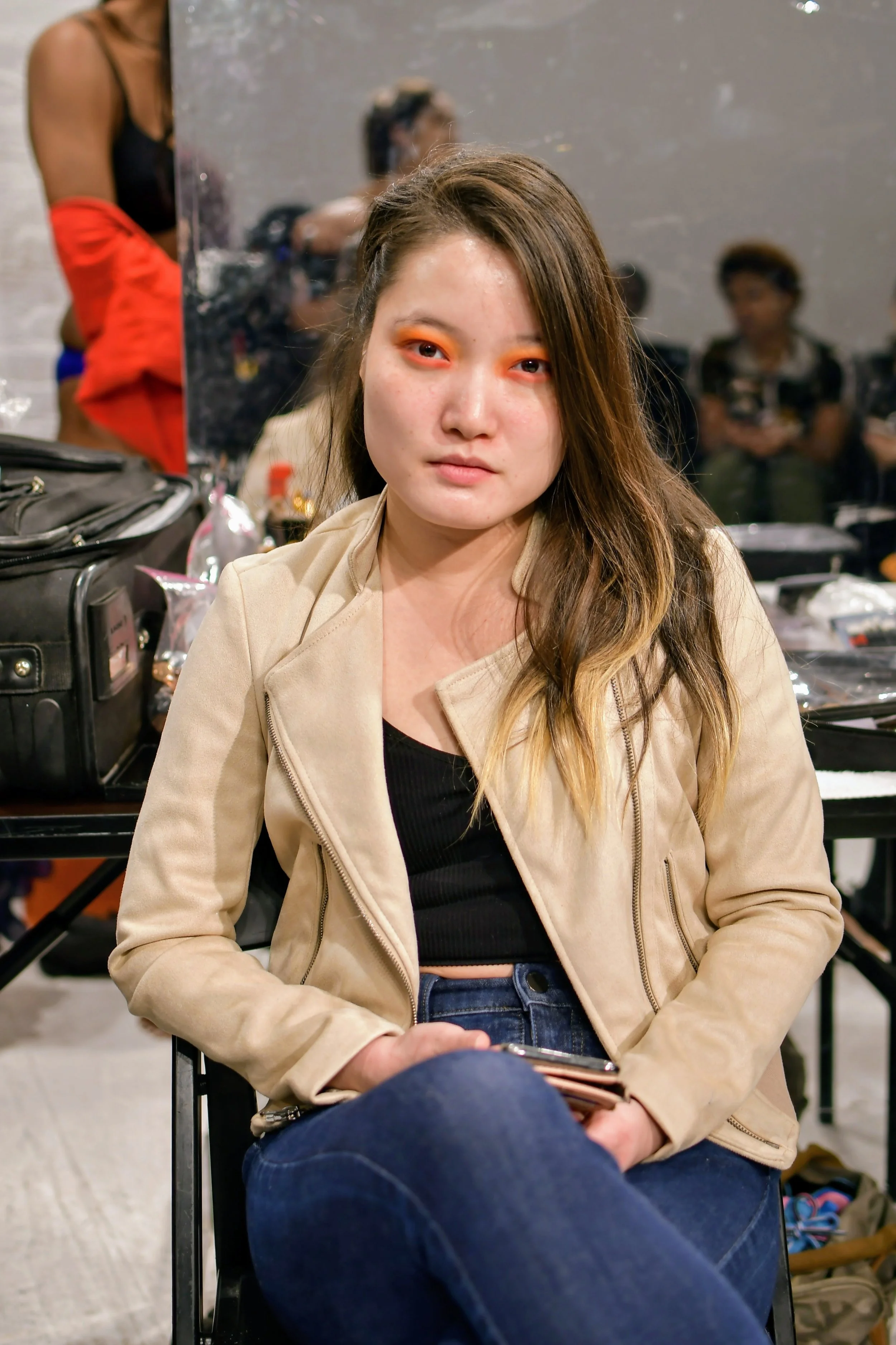 A woman sitting in front of a mirror with orange makeup on her eyelids, wearing a beige jacket and dark blue jeans, holding a small object in her hand. The background shows people and makeup stations.