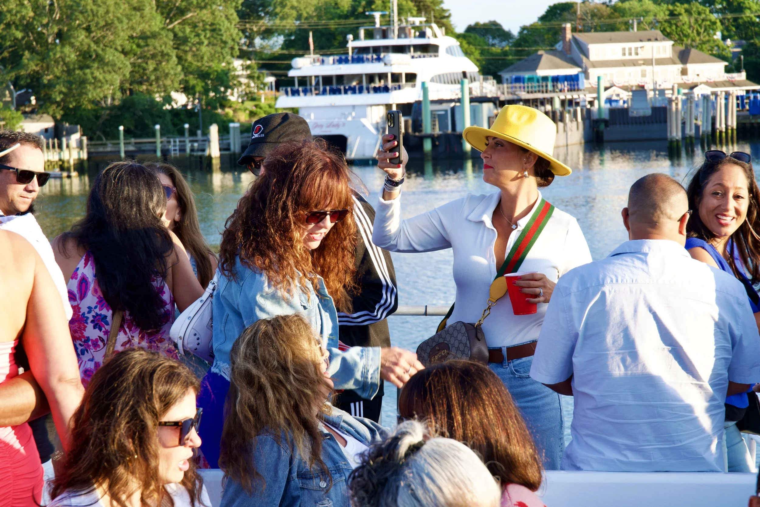 A group of people, including a woman wearing a large yellow hat, gathered outdoors near water with boats and houses in the background. Some individuals are taking photos and engaging in conversation.