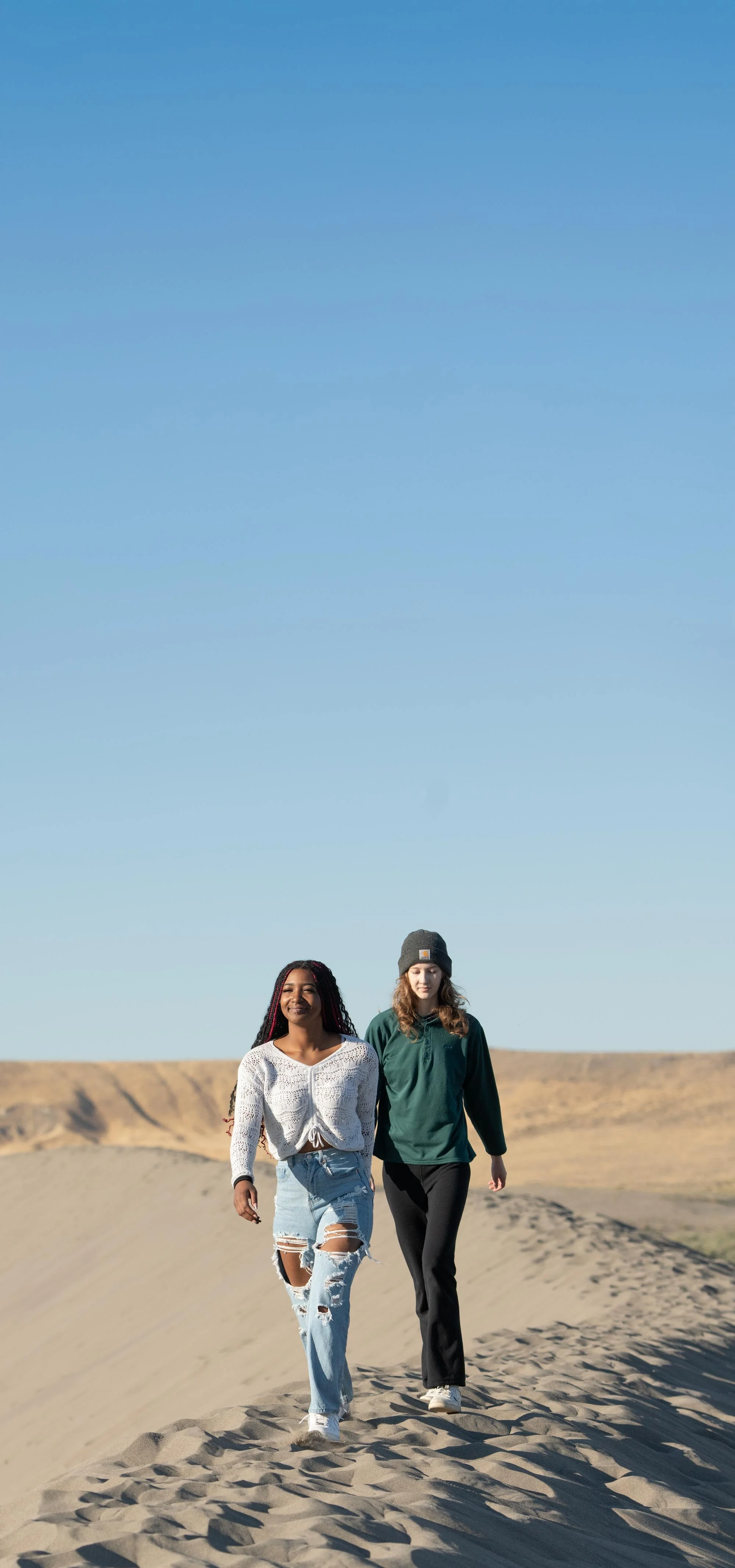 Two women walking on sand dunes in a desert with a clear blue sky.