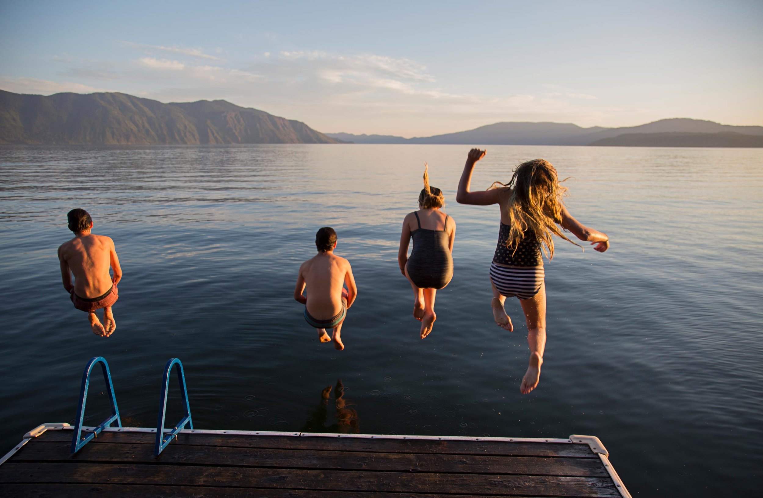 Four children jumping into a lake from a dock during sunset, with mountain scenery in the background.