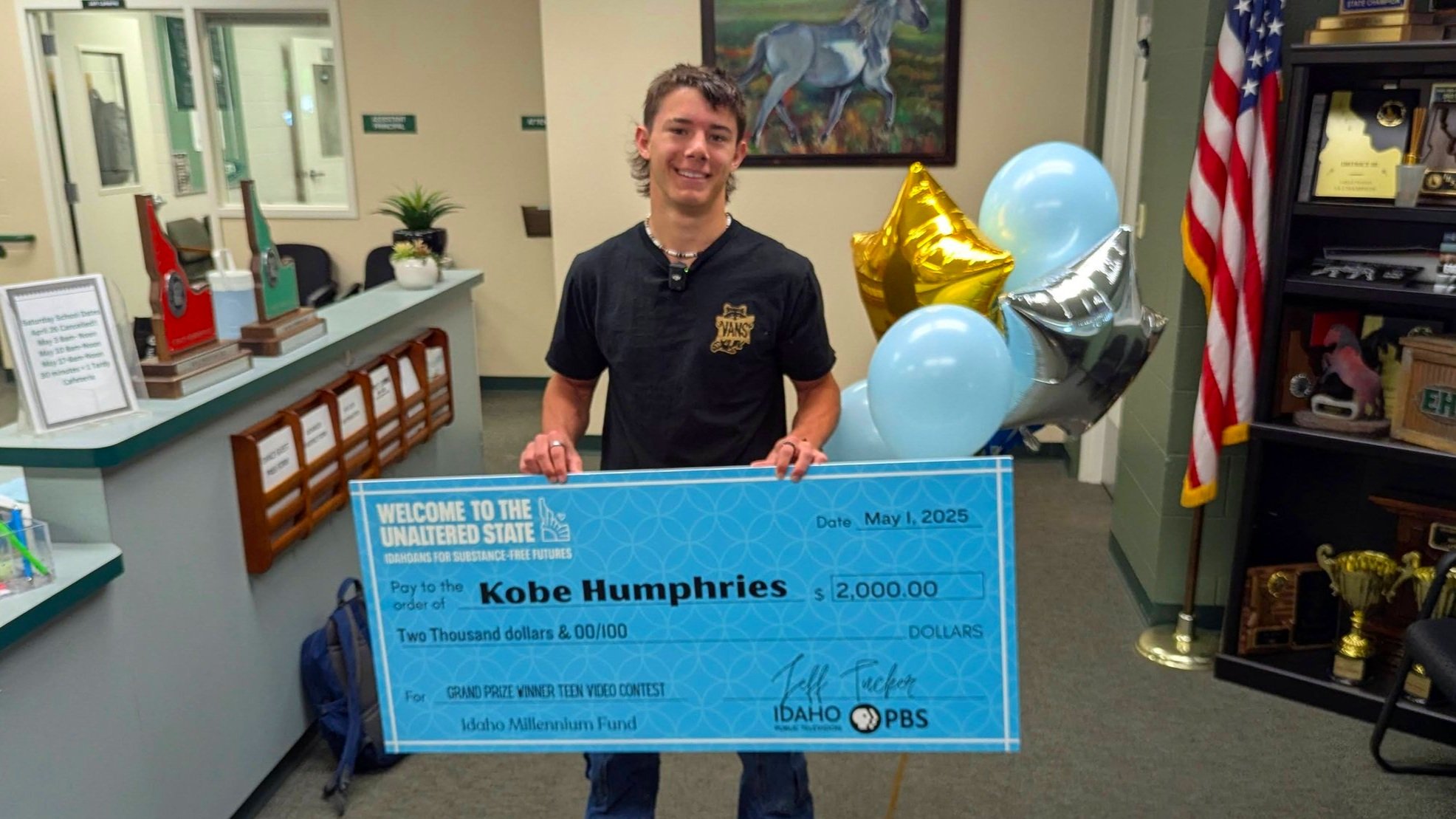 Young man smiling and holding a large check for $2,000 made out to Kobe Humphries, standing in a decorated room with balloons, an American flag, and awards displayed.