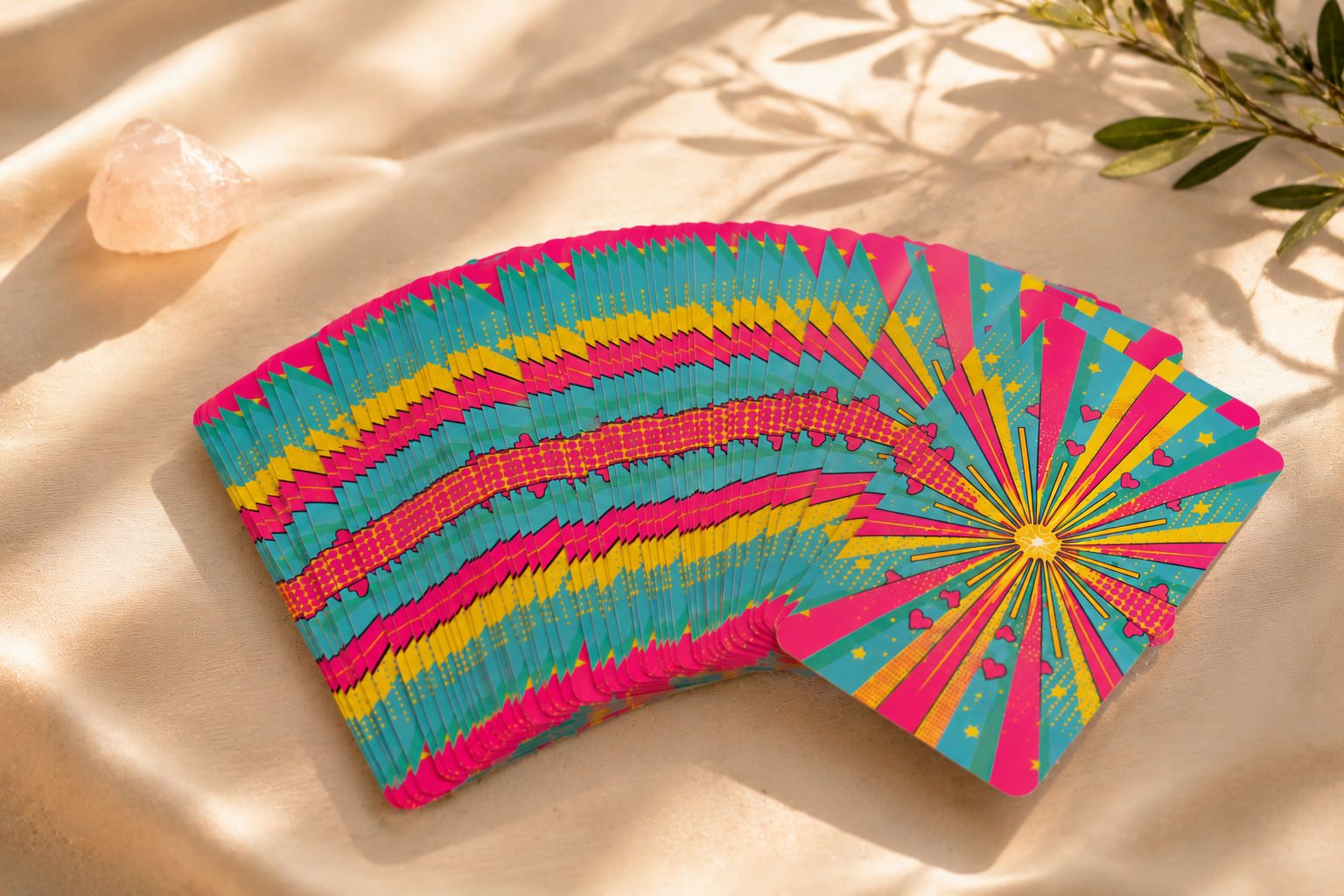 A colorful deck of playing cards with a pink, yellow, and blue design on a beige fabric surface, with some greenery and a salt rock nearby.