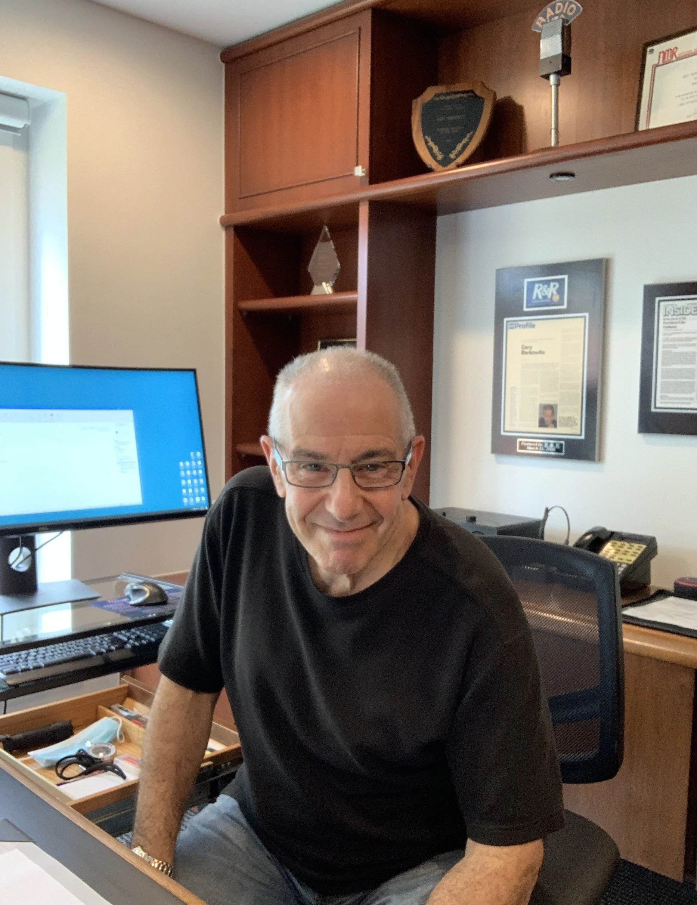 A man with short gray hair and glasses smiling at a desk in an office. He is wearing a black T-shirt and a gold watch. Behind him are framed certificates on the wall, a wooden shelf with awards and plaques, and a desktop computer with a blue screen. There are office supplies and a drawer with small items on the desk.