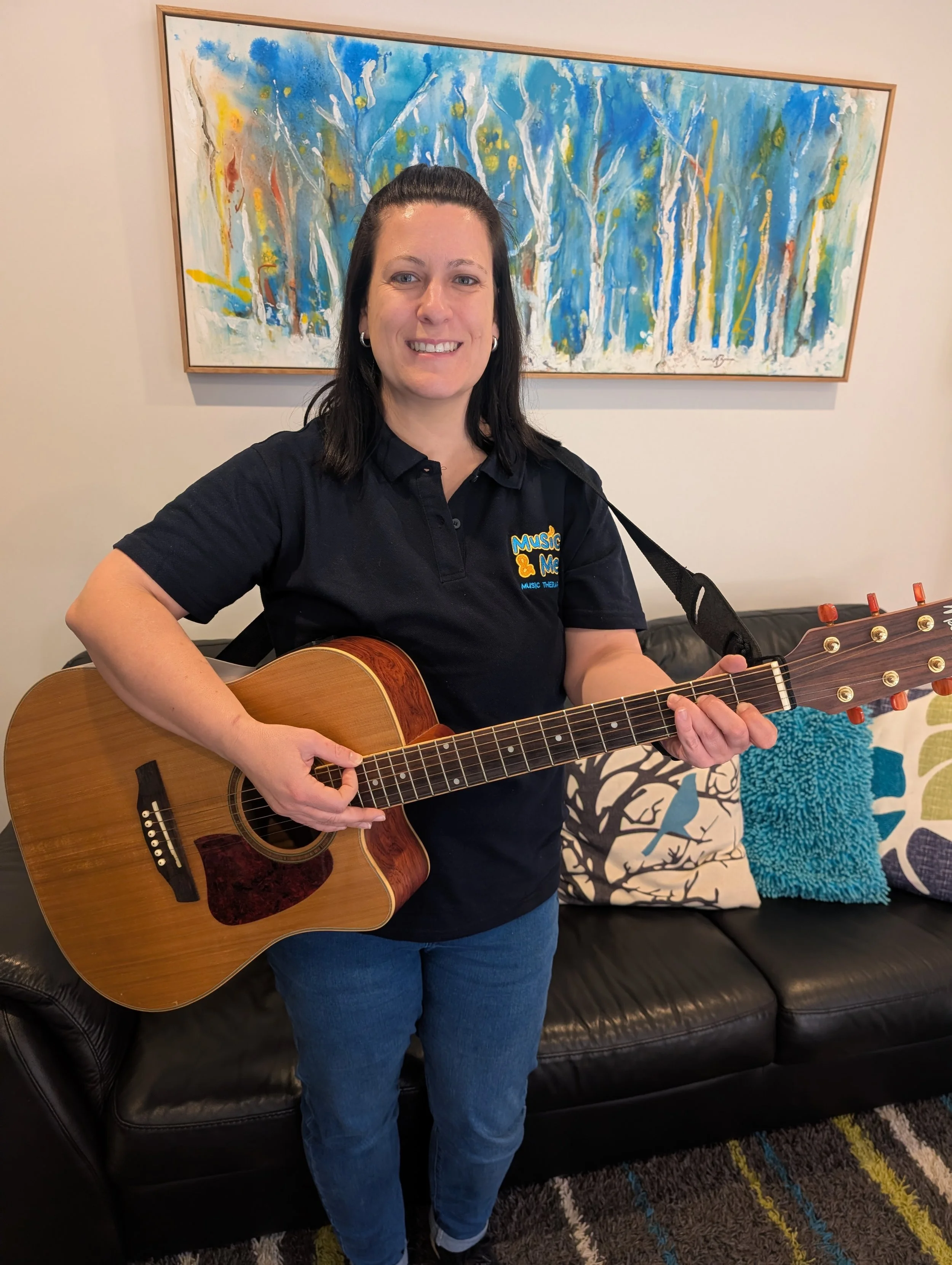A woman with dark hair, wearing a dark polo shirt with a logo that says 'Music &' and 'Music Therapy', stands in front of a colorful painting of abstract trees. She is playing an acoustic guitar and smiling. Behind her, there is a black leather sofa with decorative pillows, including a blue textured pillow, in a room with a patterned rug.
