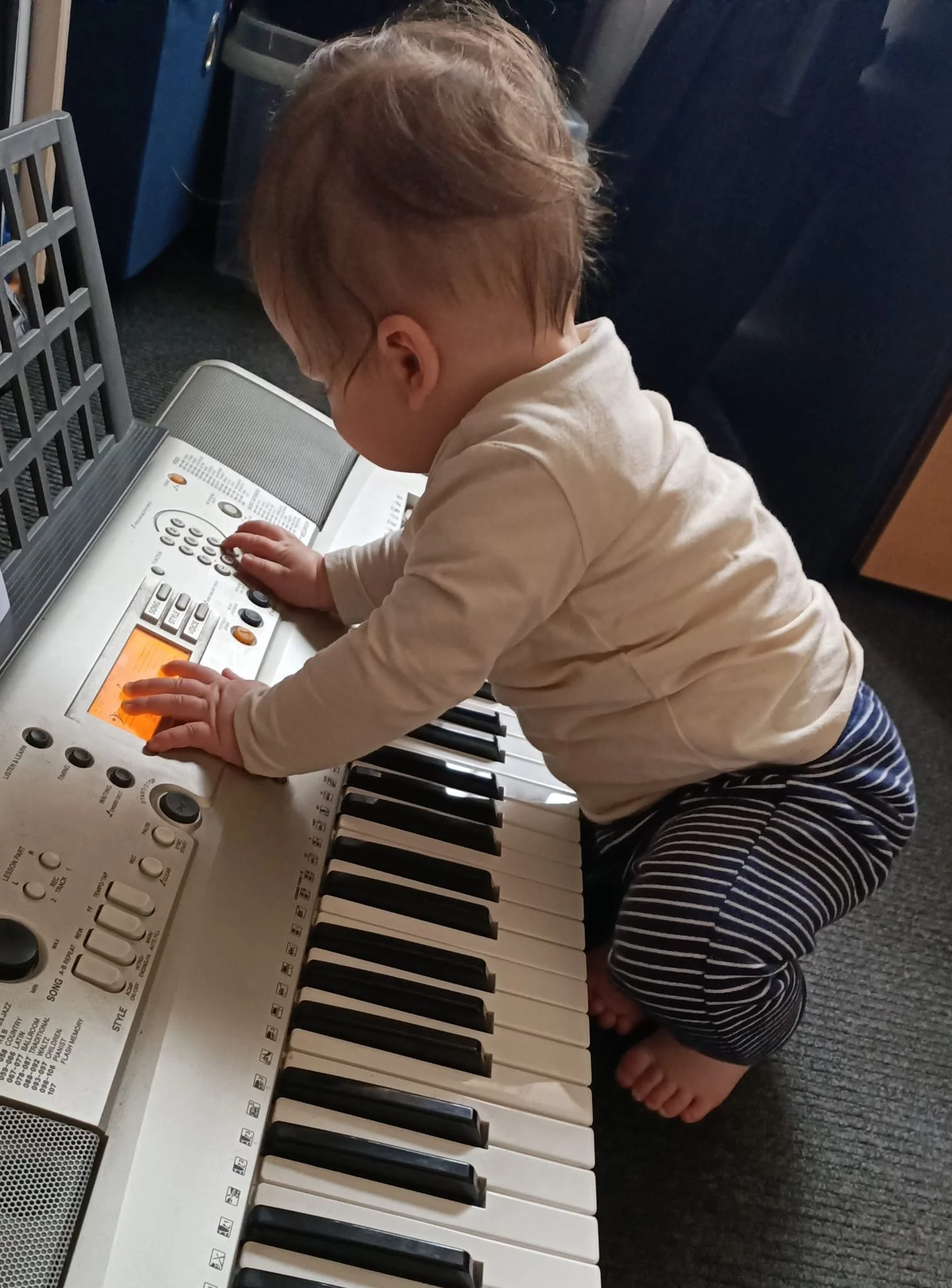 A young child with short light brown hair, kneeling on the carpeted floor, dressed in a cream long sleeve shirt and striped pants, playing a small keyboard or synthesizer. The child appears to be pressing the keys or buttons.