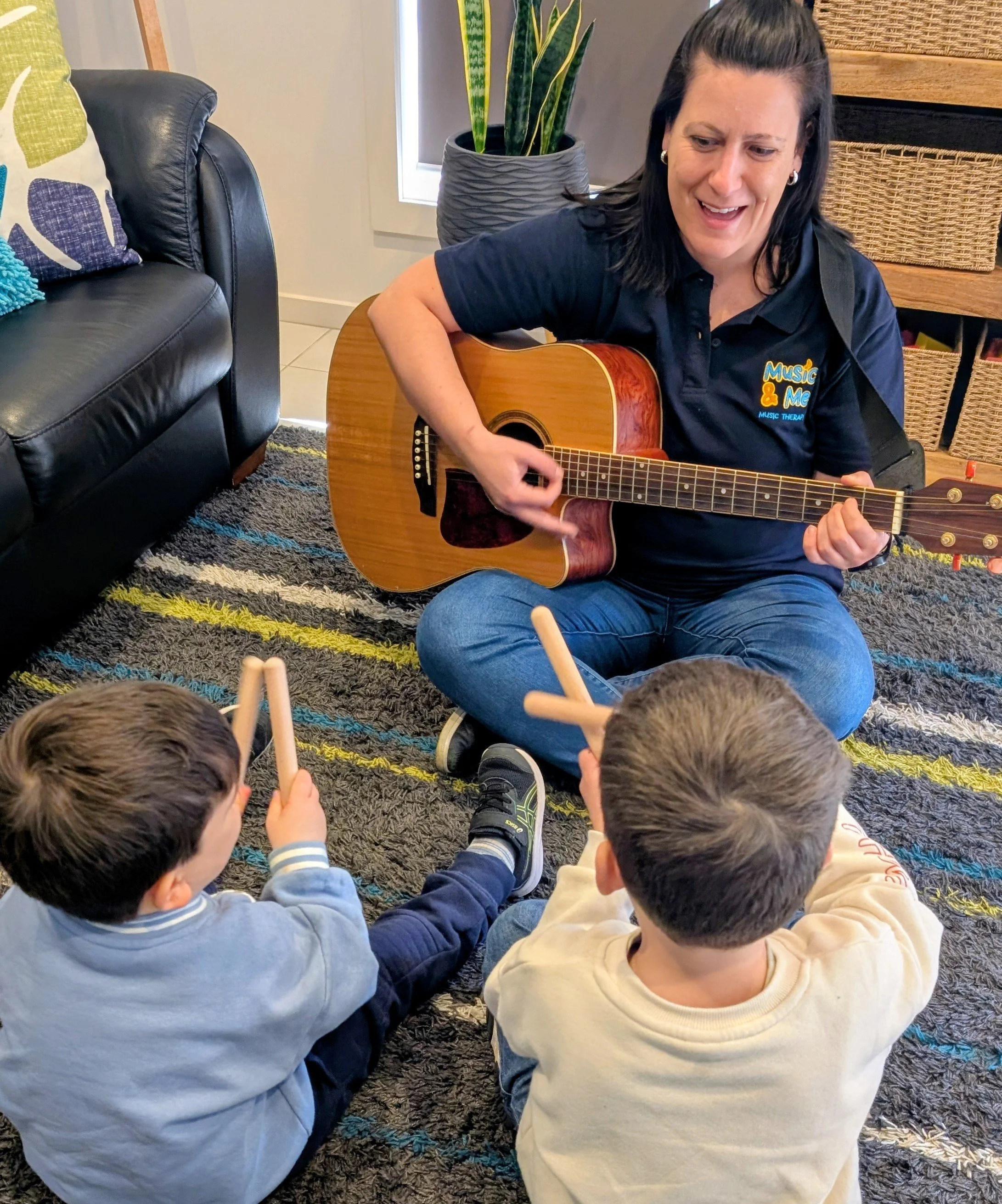 A woman playing an acoustic guitar surrounded by children sitting on a colorful rug in a cozy room with a black leather chair, a potted plant, and woven baskets in the background.