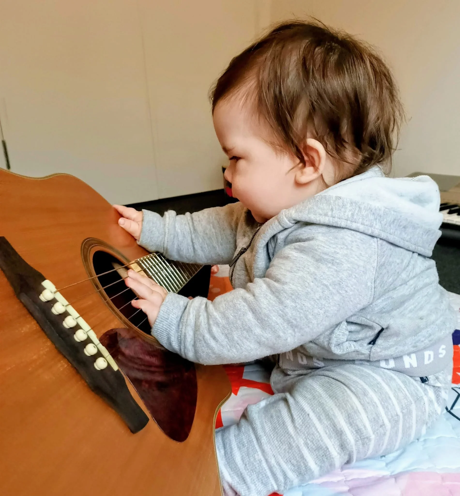 A young child playing a brown acoustic guitar at a table, wearing a grey hoodie.