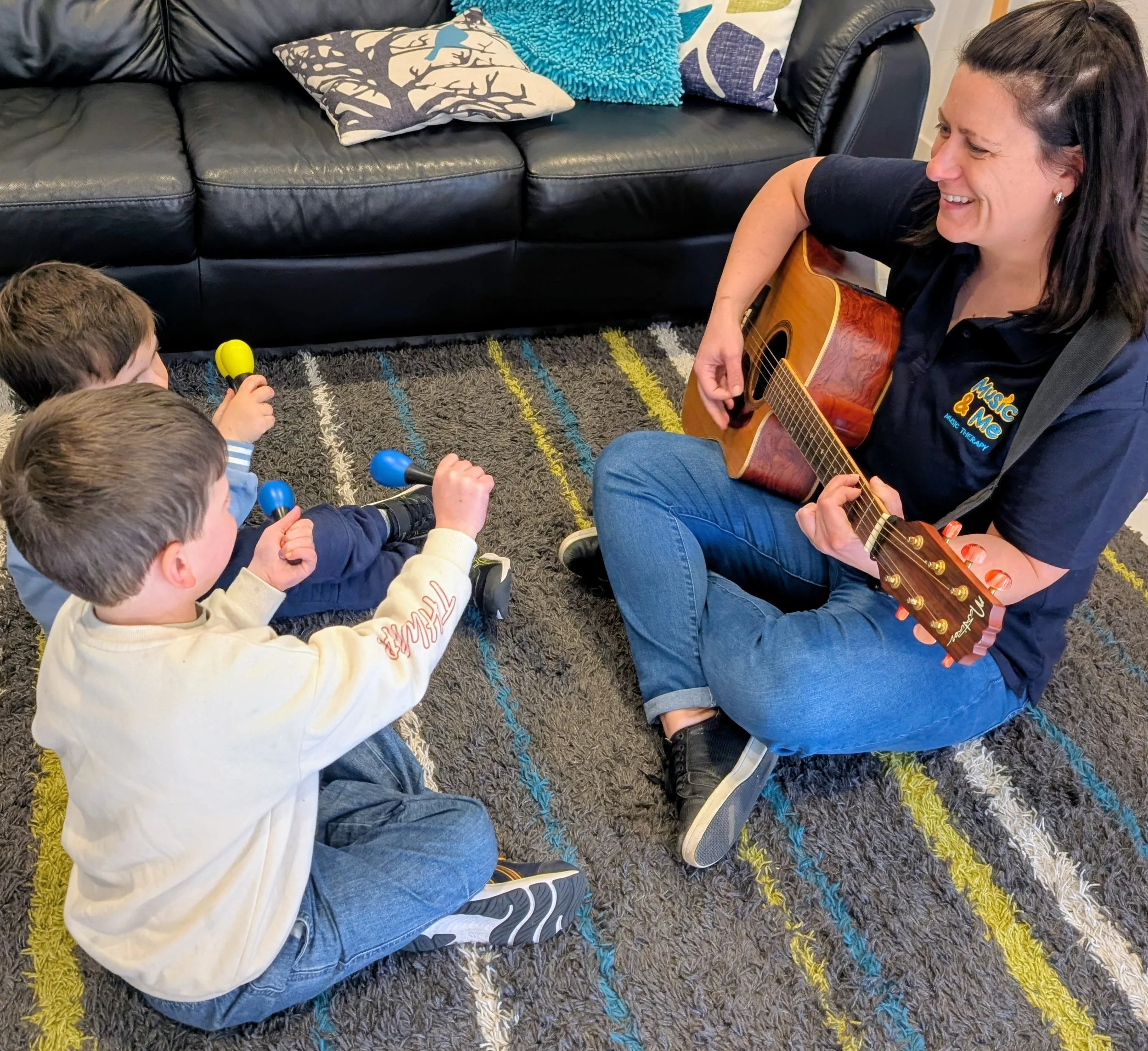 A woman playing guitar to a group of children sitting on a colorful rug, indoors with a black leather couch and decorative pillows in the background.