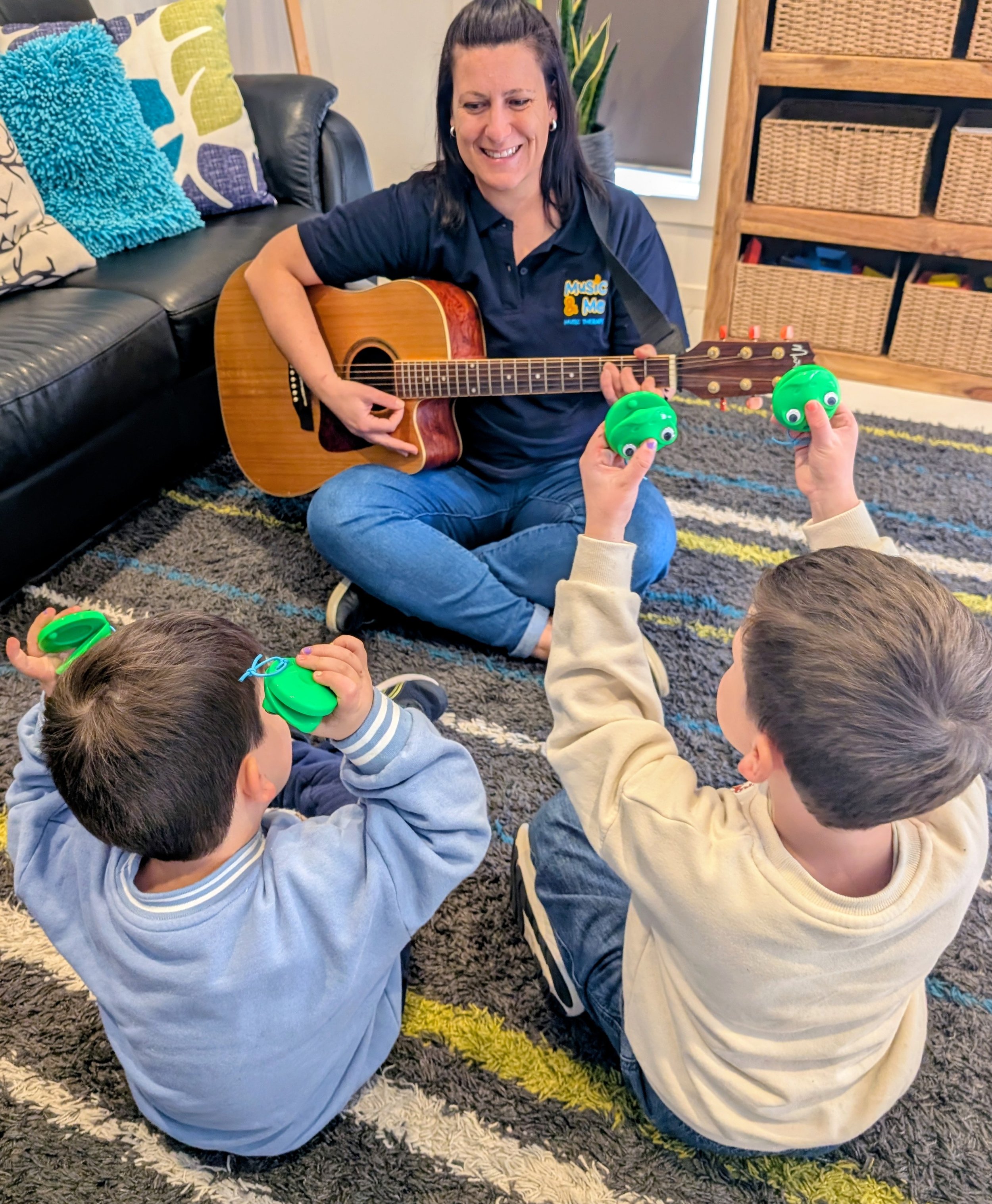 A woman playing guitar while children sitting on the floor hold green toy frogs with googly eyes. The woman is smiling and sitting on a rug in a cozy indoor setting with cushions and a wooden shelf in the background.