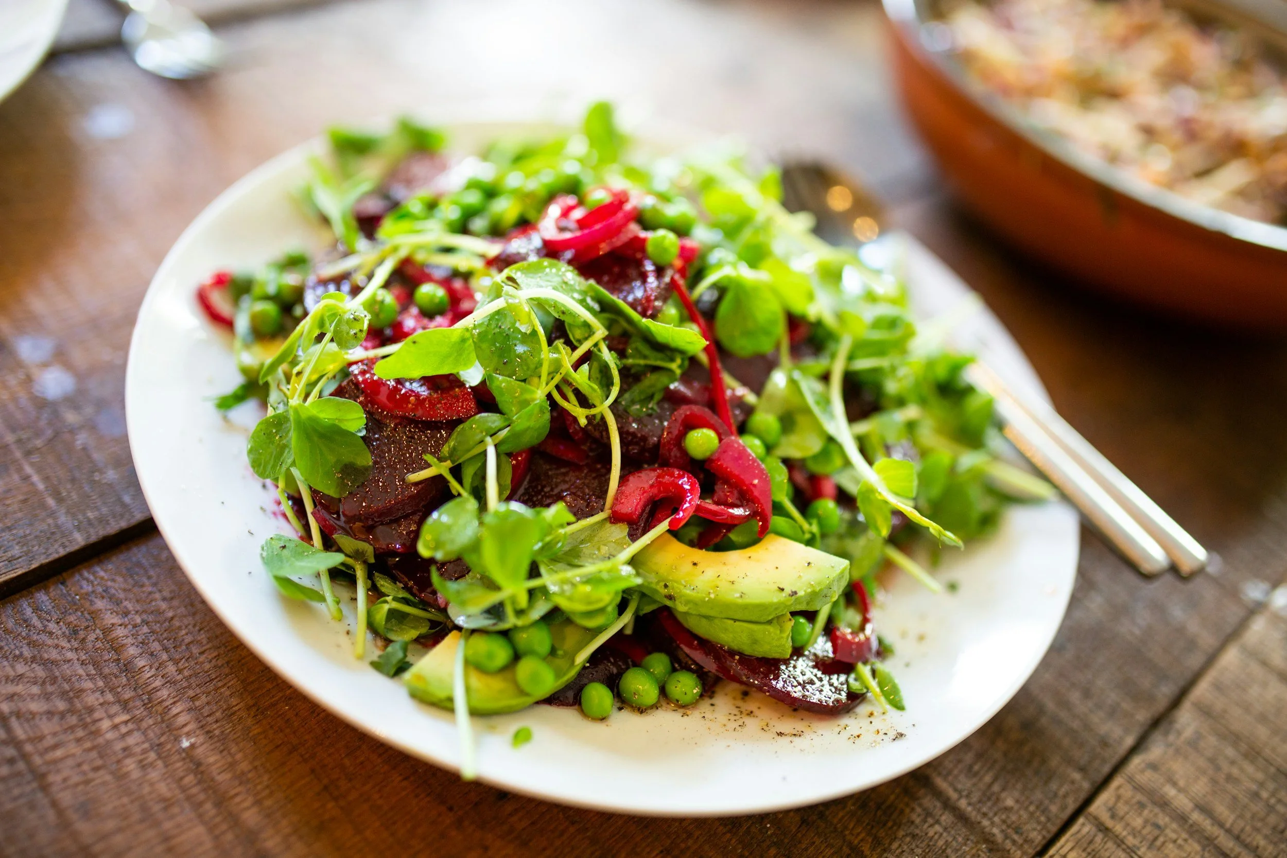 A white plate of salad with greens, peas, sliced beets, and avocado on a wooden table, with a baked dish in the background.