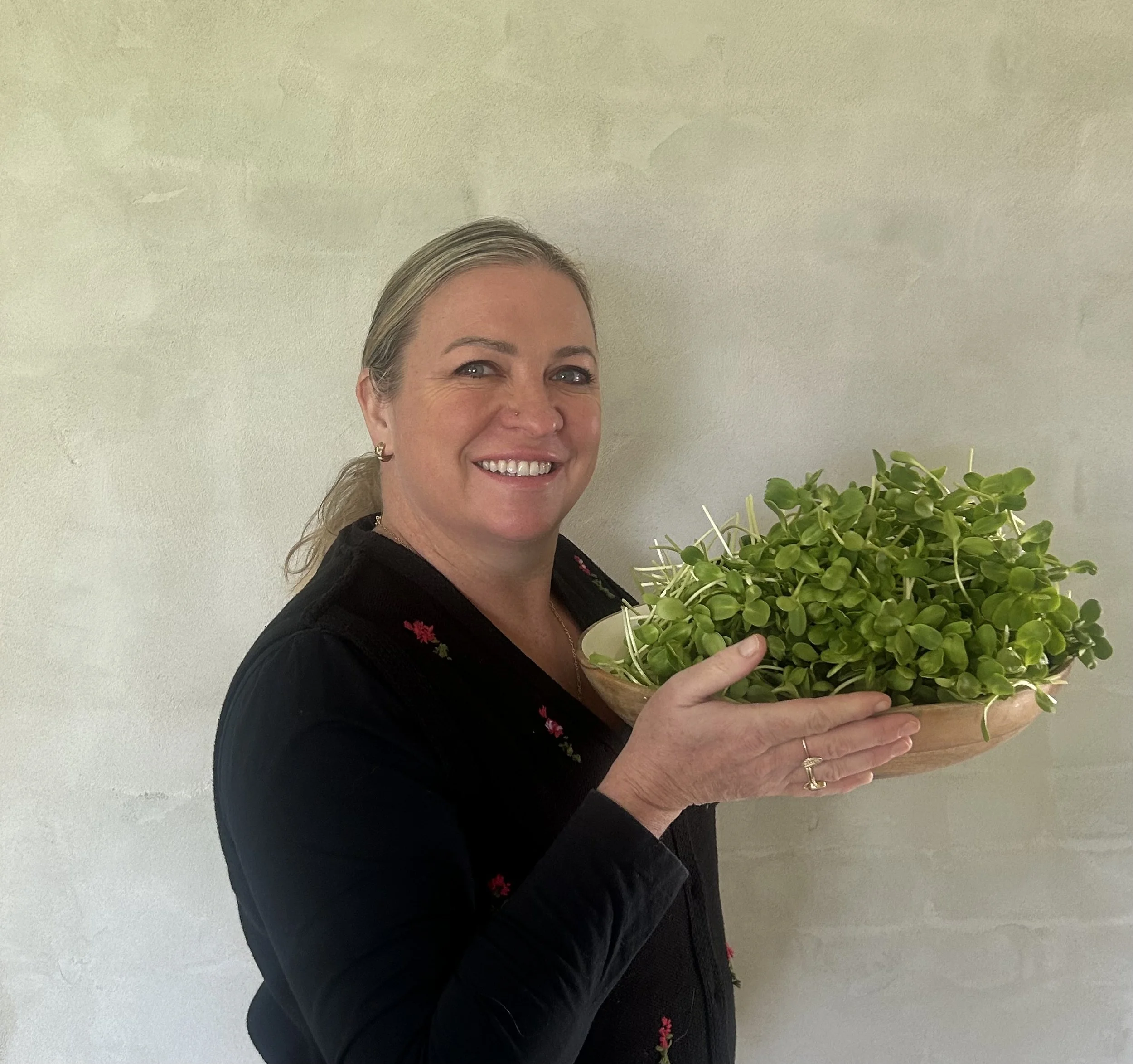 A smiling woman with blonde hair holding a large green potted plant in front of a plain light-colored wall.