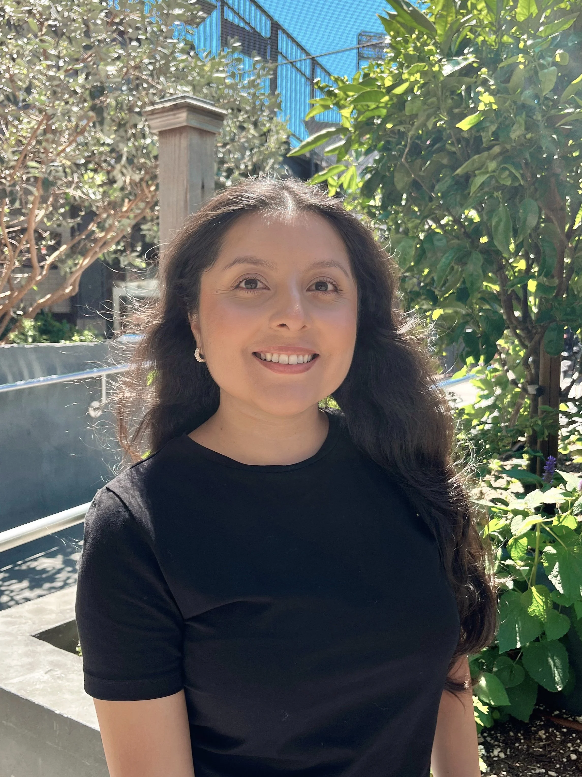 A woman with long, dark hair smiling outdoors in sunlight, wearing a black t-shirt, with greenery and bushes in the background.