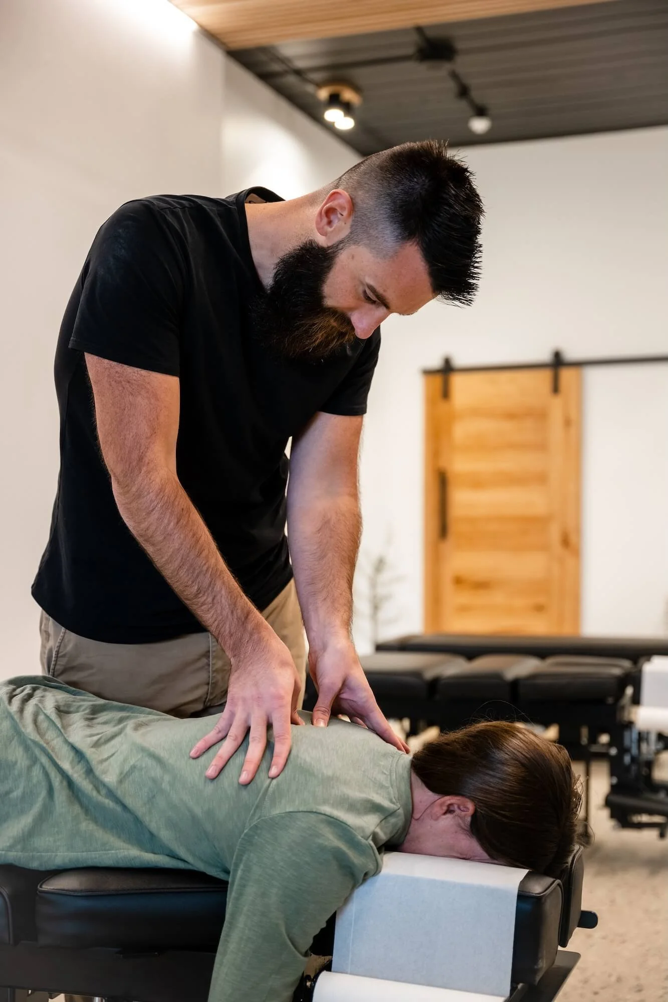 A man with a beard and dark hair is performing a chiropractic adjustment or massage on a woman lying face down on a massage table. The woman has brown hair and is wearing a green shirt. The setting appears to be a wellness or chiropractic clinic with a minimalist decor, including a white wall, black chairs, and a sliding barn door in the background.